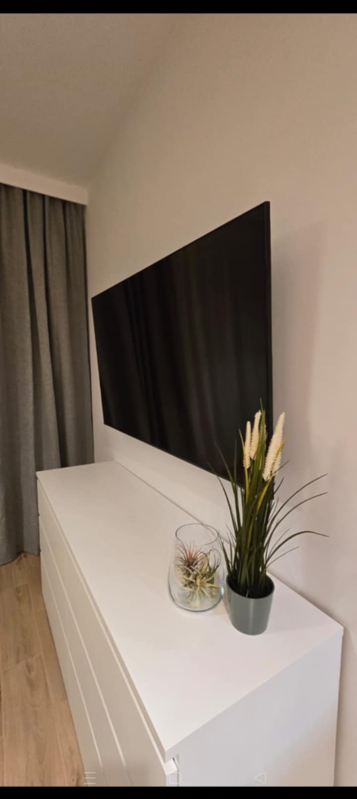 Nowoczesny salon: Telewizor na ścianie nad białą komodą, obok zasłony. Dekoracje: szklany wazon z rośliną i wysoka trawa w doniczce. Minimalistyczny design.