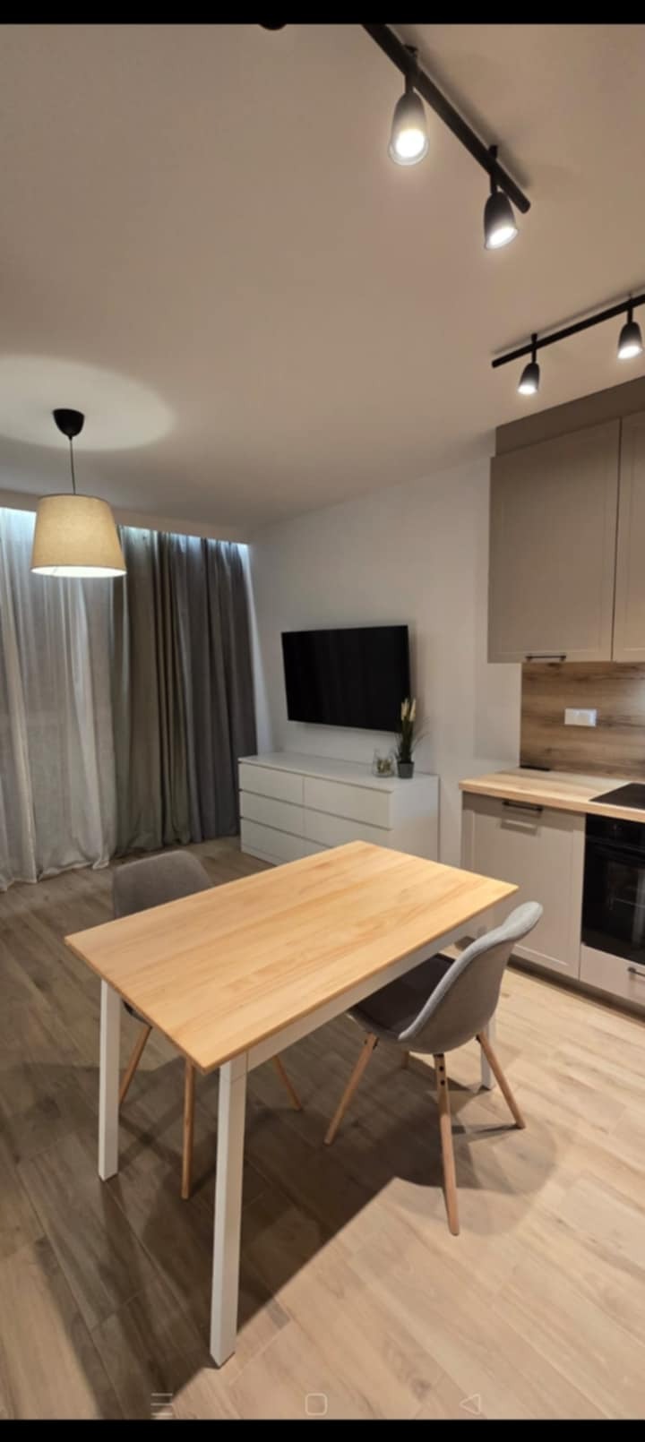 Jasne wnętrze mieszkania z drewnianym stołem i szarą tapicerowaną krzesłem. W tle komoda, telewizor i zasłony. Oświetlenie punktowe na suficie.