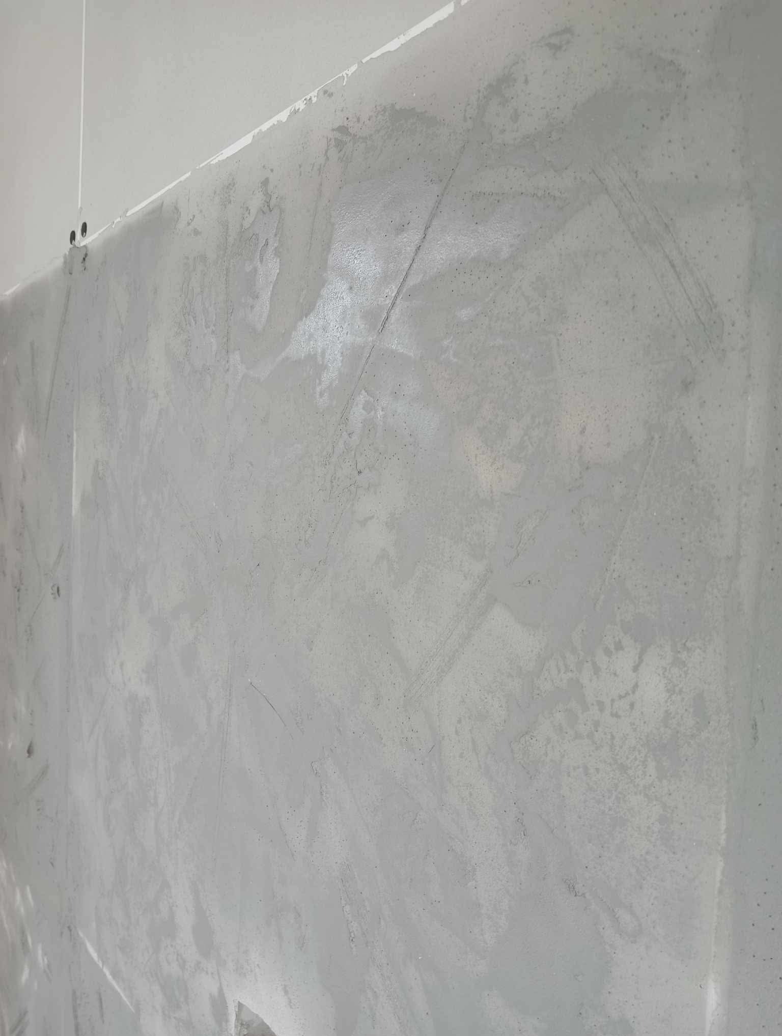 Świeżo nałożona, szara gładź szpachlowa na ścianie, widoczne ślady po szpachelce. Ujęcie z dołu, podkreślające fakturę i nierówności. Oświetlenie naturalne, dzienne.
