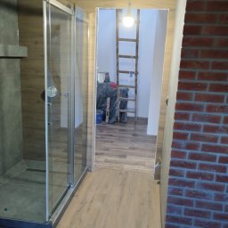 Milpa - Nowoczesna kabina prysznicowa z przeszklonymi drzwiami, wykończona płytkami imitującymi drewno. W tle drabina i materiały budowlane, ściana z cegły.