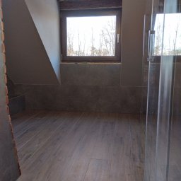 Milpa - Ukończona łazienka na poddaszu z drewnianą podłogą i oknem. Widoczna kabina prysznicowa ze szkła. Ściany wykończone płytkami w odcieniach szarości.