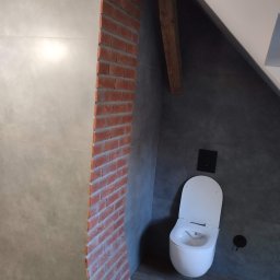 Milpa - Wykończona łazienka w szarościach z akcentem ceglanej ściany. Uwagę przykuwa nowoczesna toaleta i drewniany element konstrukcyjny pod skosem.