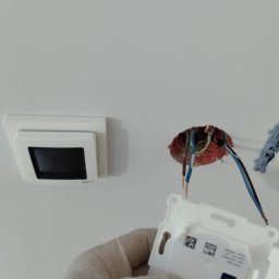 Milpa - Montaż panelu sterowania ogrzewaniem podłogowym: widoczne przewody, puszka instalacyjna i dłoń w rękawicy trzymająca element panelu. Biała ściana w tle.