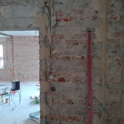 Milpa - Ceglana ściana w trakcie remontu z poziomnicą, widoczne ślady po instalacji elektrycznej i przygotowania do gładzi. W tle widok na odnowione pomieszczenie.