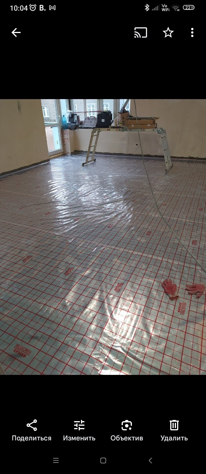 Przygotowanie do montażu ogrzewania podłogowego: folia refleksyjna z siatką, drabina i narzędzia w jasnym wnętrzu. Widoczne rękawice robocze.