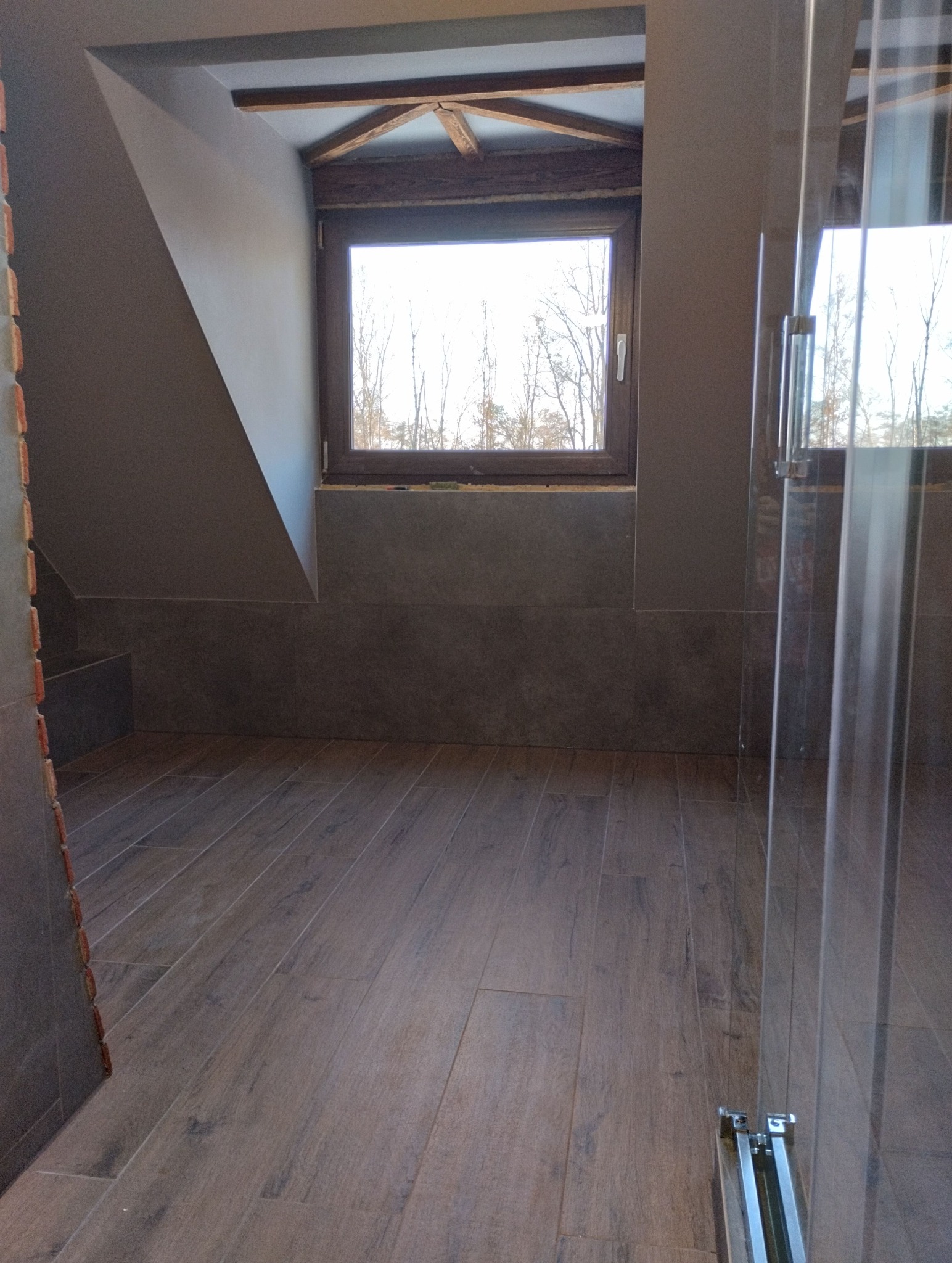 Ukończona łazienka na poddaszu z drewnianą podłogą i oknem. Widoczna kabina prysznicowa ze szkła. Ściany wykończone płytkami w odcieniach szarości.