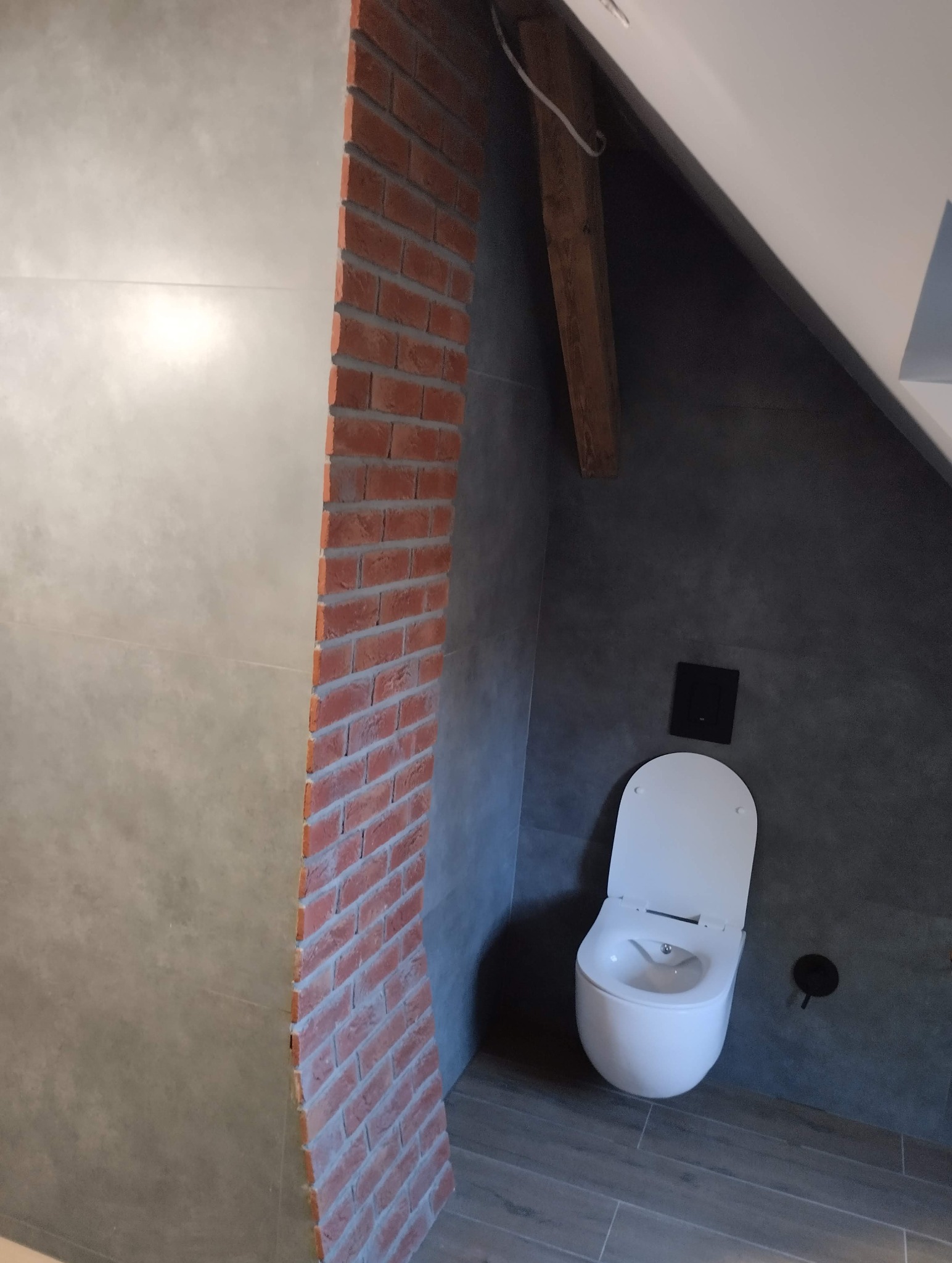 Wykończona łazienka w szarościach z akcentem ceglanej ściany. Uwagę przykuwa nowoczesna toaleta i drewniany element konstrukcyjny pod skosem.