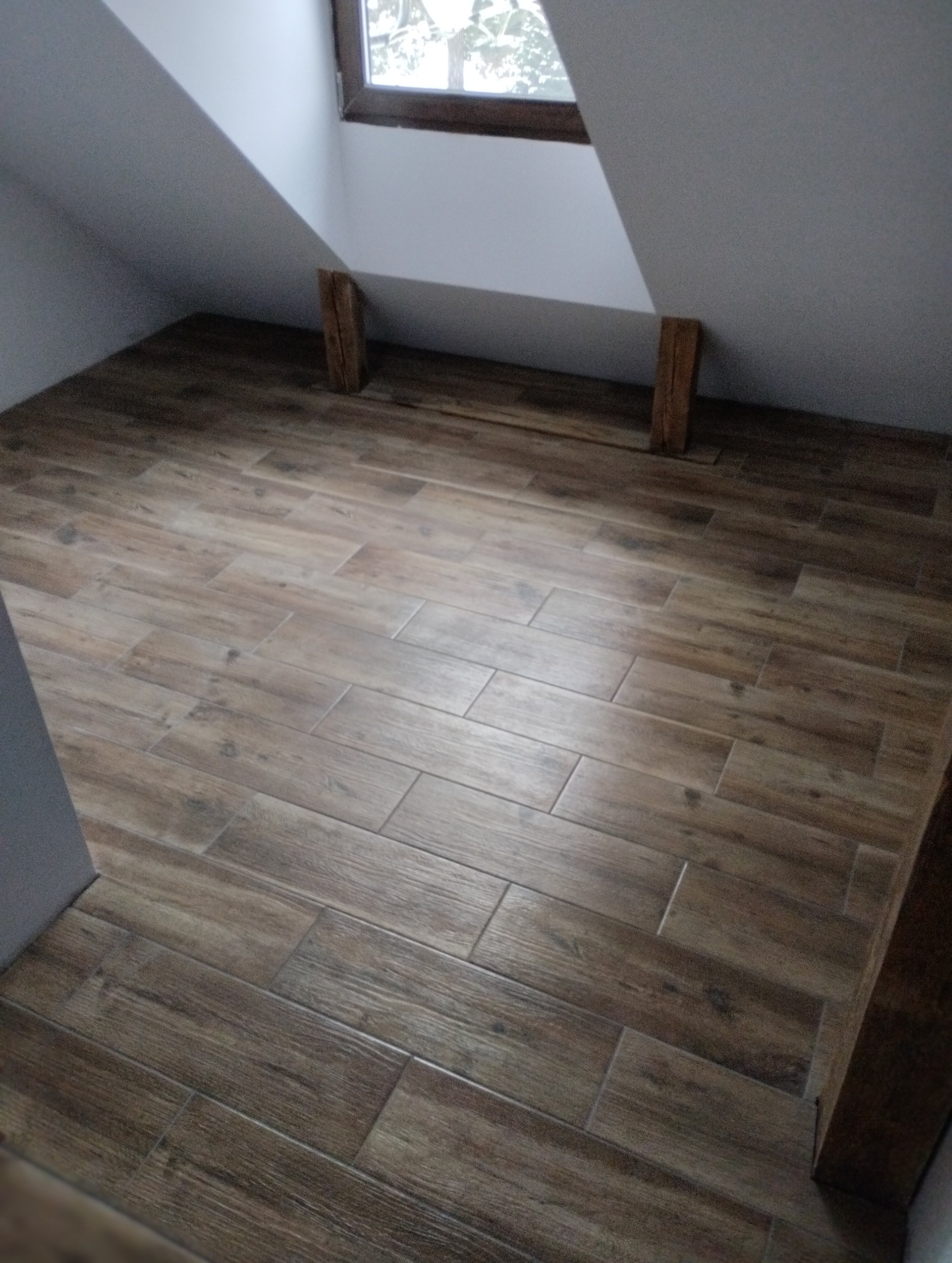 Ukończona podłoga z płytek imitujących drewno w pokoju na poddaszu, z oknem i drewnianymi elementami konstrukcyjnymi. Widok z góry, ciepłe oświetlenie.