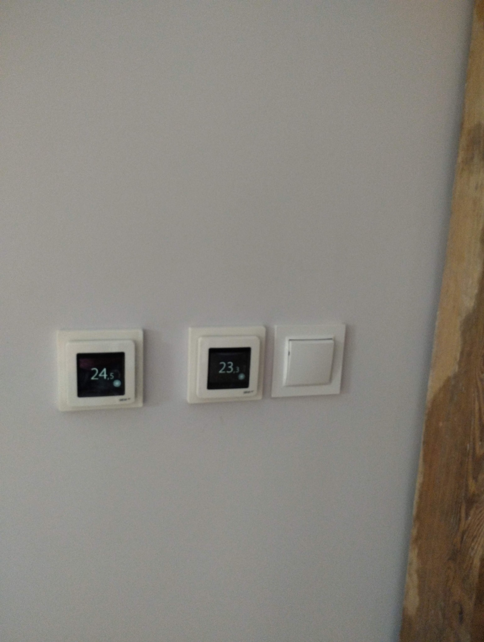 Szara ściana z dwoma termostatami pokazującymi temperaturę 24.5 i 23.3 stopni Celsjusza oraz włącznikiem światła. Fragment drewnianej deski po prawej stronie.