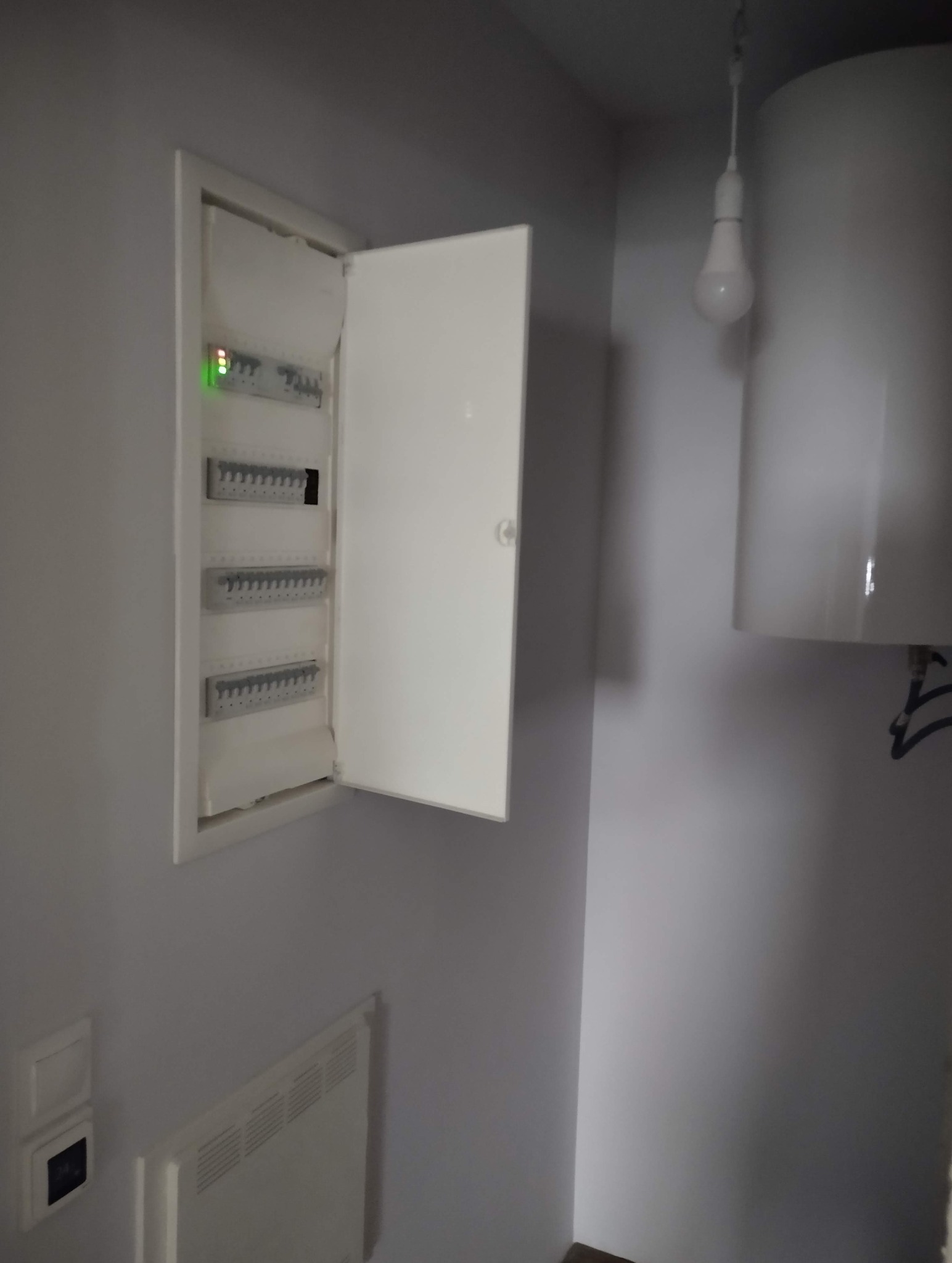 Otwarta skrzynka elektryczna z widocznymi bezpiecznikami na szarej ścianie, obok biały bojler i żarówka. Widoczny fragment grzejnika elektrycznego.