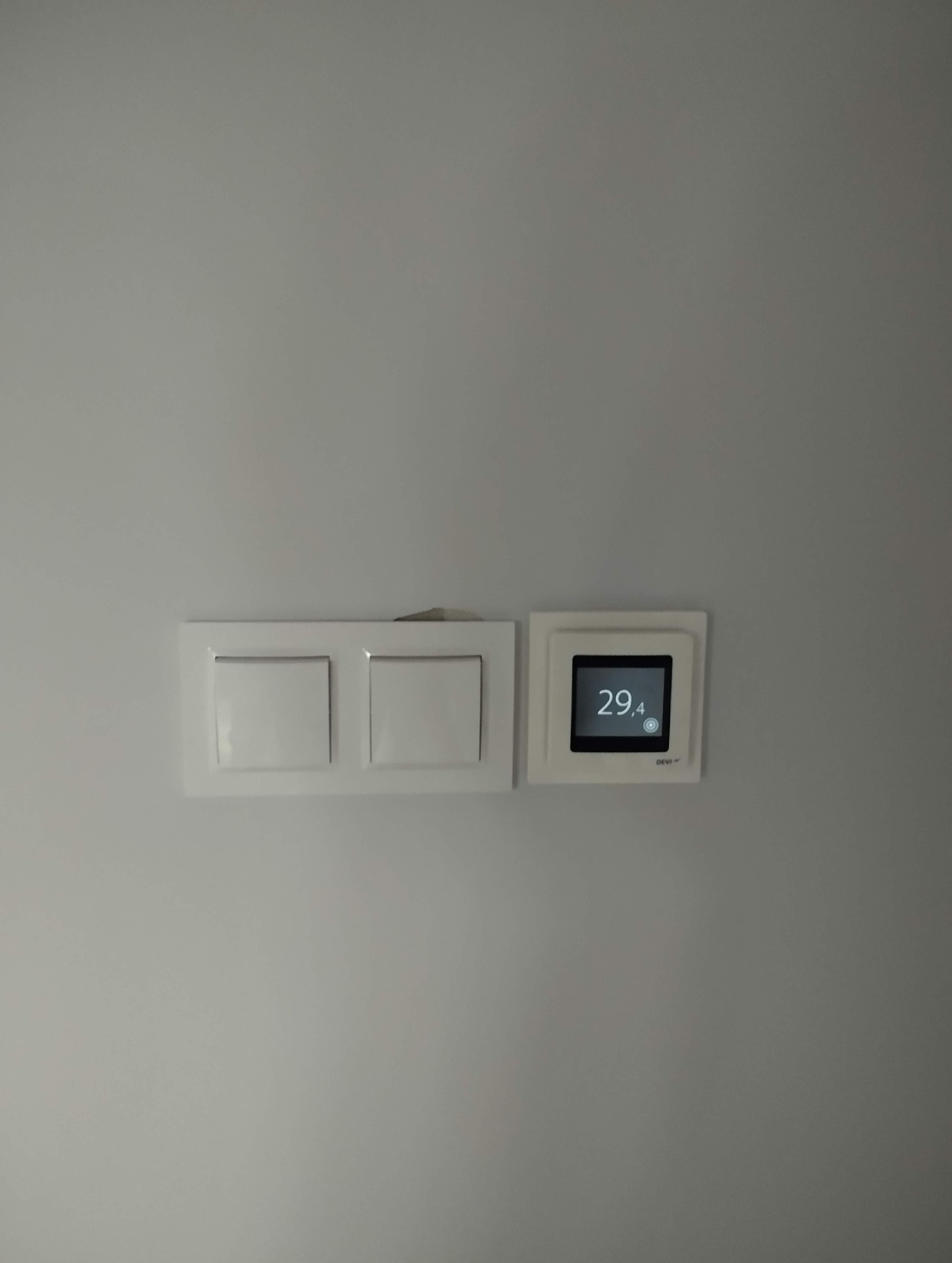 Biała ściana z zamontowanym panelem sterowania ogrzewaniem podłogowym marki Devi, obok włącznik światła. Minimalistyczny design.