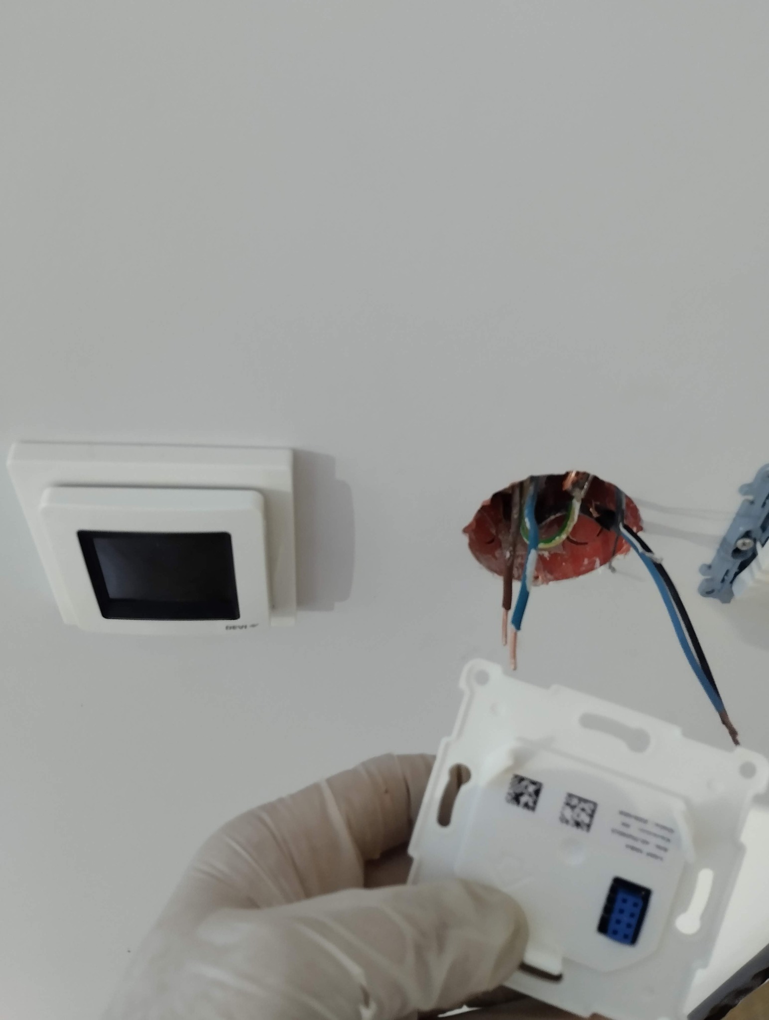 Montaż panelu sterowania ogrzewaniem podłogowym: widoczne przewody, puszka instalacyjna i dłoń w rękawicy trzymająca element panelu. Biała ściana w tle.