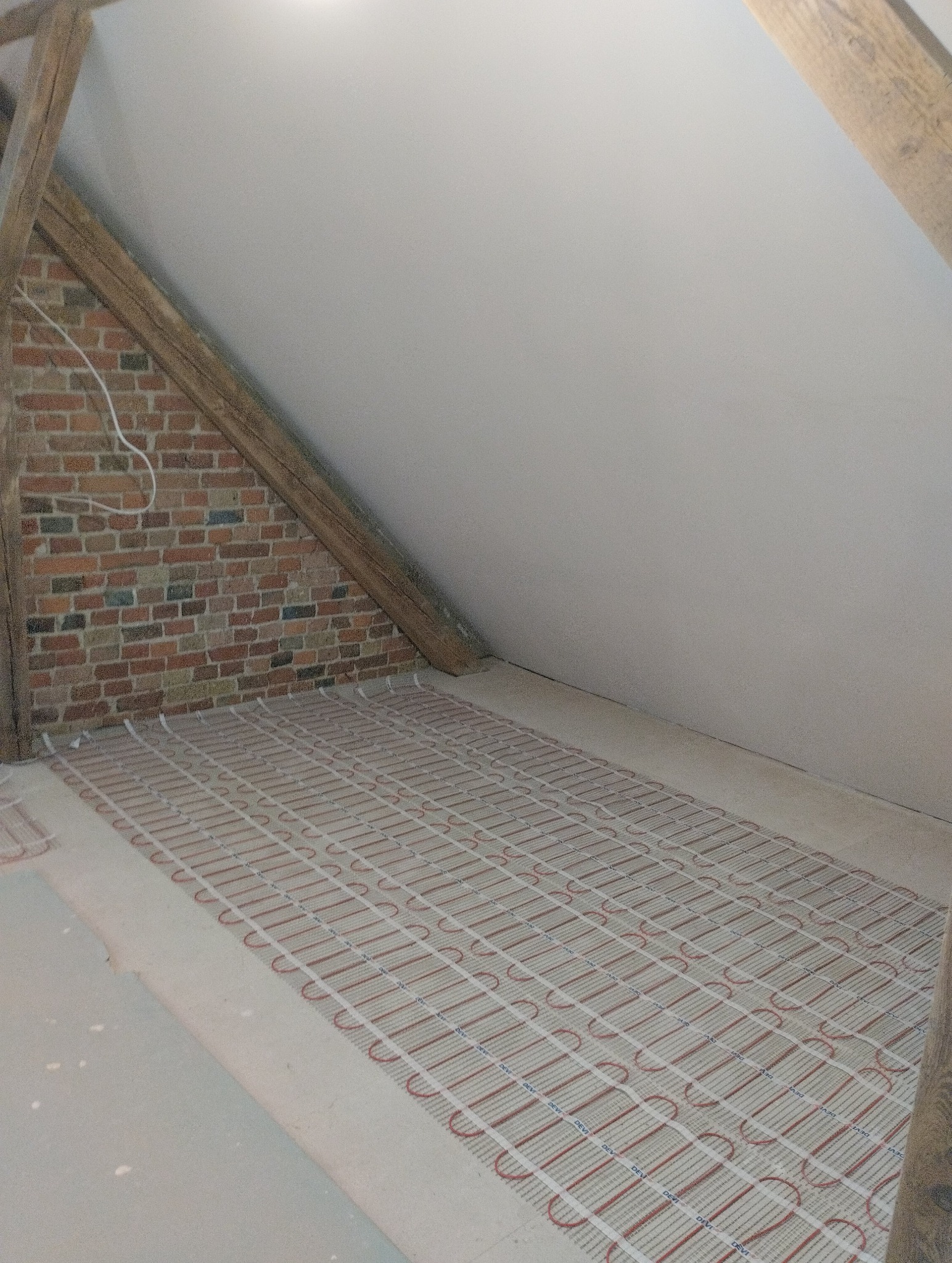 Podłogówka elektryczna Devi ułożona na poddaszu, widok z góry. Czerwone kable grzewcze na białej siatce, ceglana ściana w tle, drewniana belka stropowa.