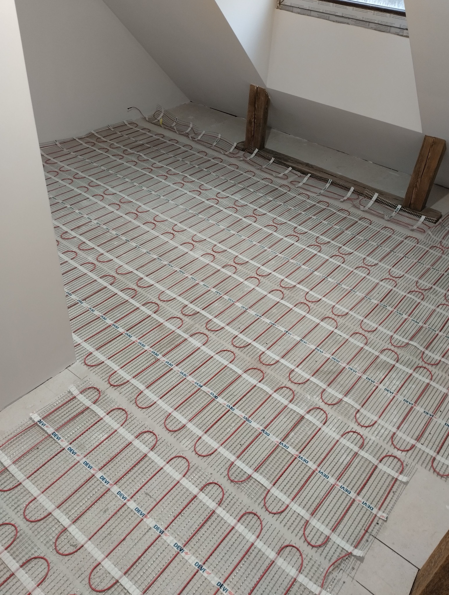 Instalacja ogrzewania podłogowego DEVI w poddaszu, widoczne kable grzewcze ułożone na podłożu, przygotowane do zalania wylewką. Elementy drewniane w tle.