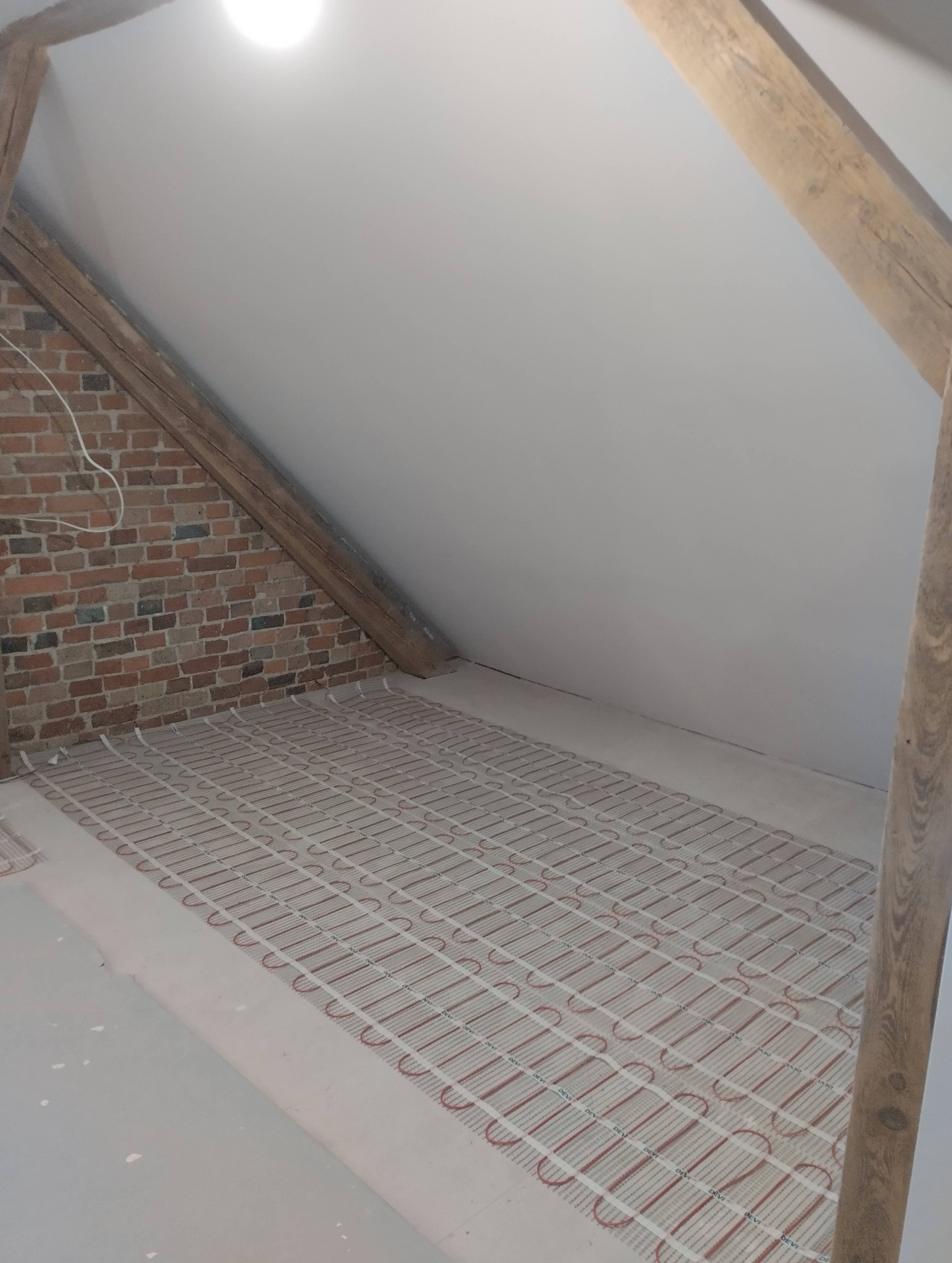 Podłogówka elektryczna w poddaszu z ceglaną ścianą i drewnianymi belkami. Mata grzewcza ułożona na podłożu, gotowa do zalania wylewką. Oświetlenie sufitowe.