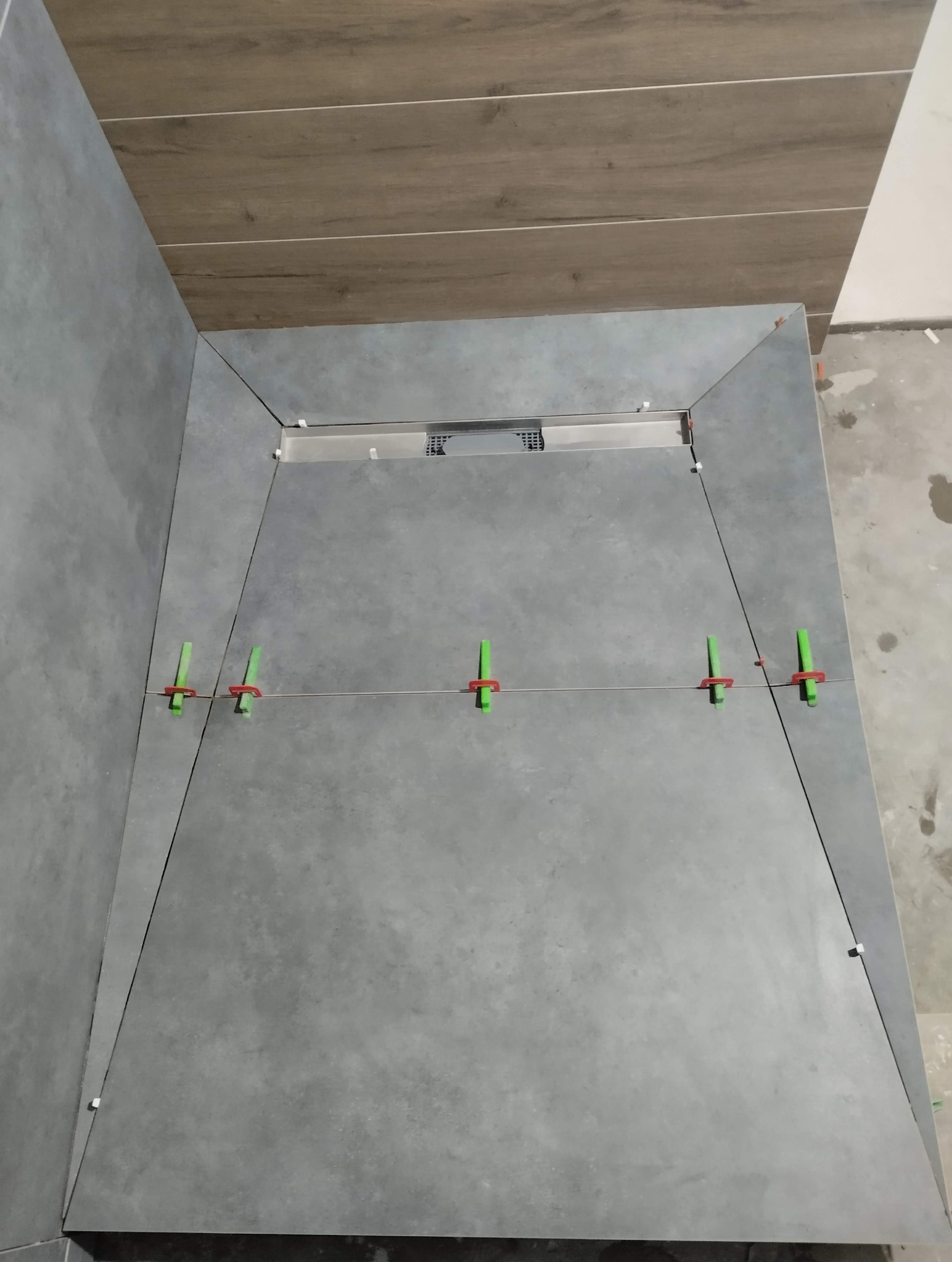 Szare płytki podłogowe w kabinie prysznicowej z odpływem liniowym, wyrównane za pomocą klipsów i sznurka. Widoczne płytki na ścianach i drewnopodobne na suficie.