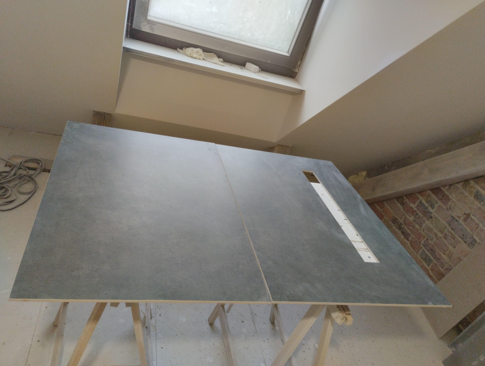 Dwie duże, szare płyty ułożone na drewnianych kozłach w pomieszczeniu w trakcie prac wykończeniowych. Widoczne okno dachowe i fragment ściany z cegły.