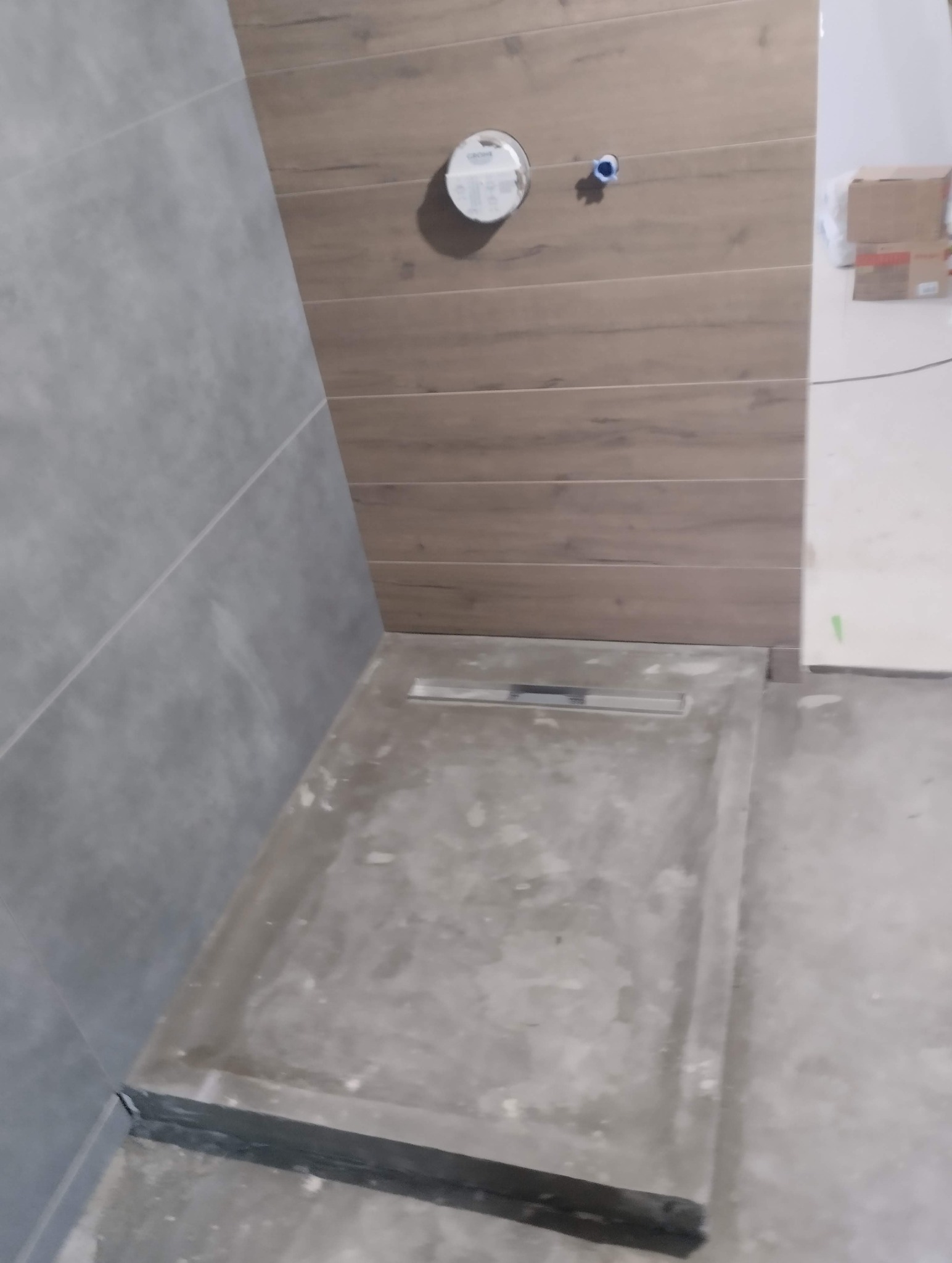 Brodzik z odpływem liniowym w trakcie montażu, szare płytki na ścianie, imitacja drewna na górze. Widoczne elementy instalacji wodnej.