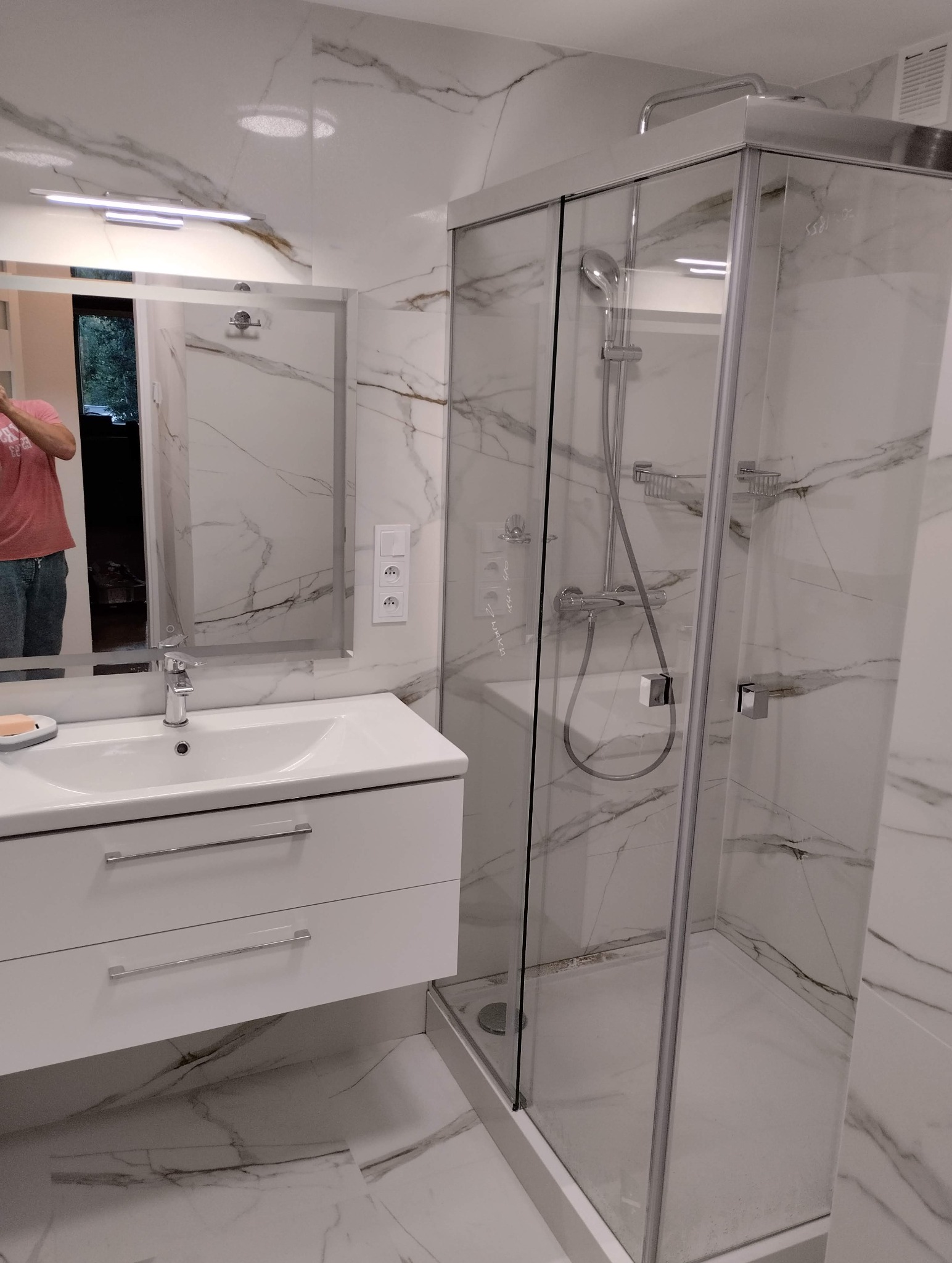 Biała łazienka z kabiną prysznicową i umywalką na szafce, wykończona płytkami imitującymi marmur. Widoczne detale armatury i oświetlenia.