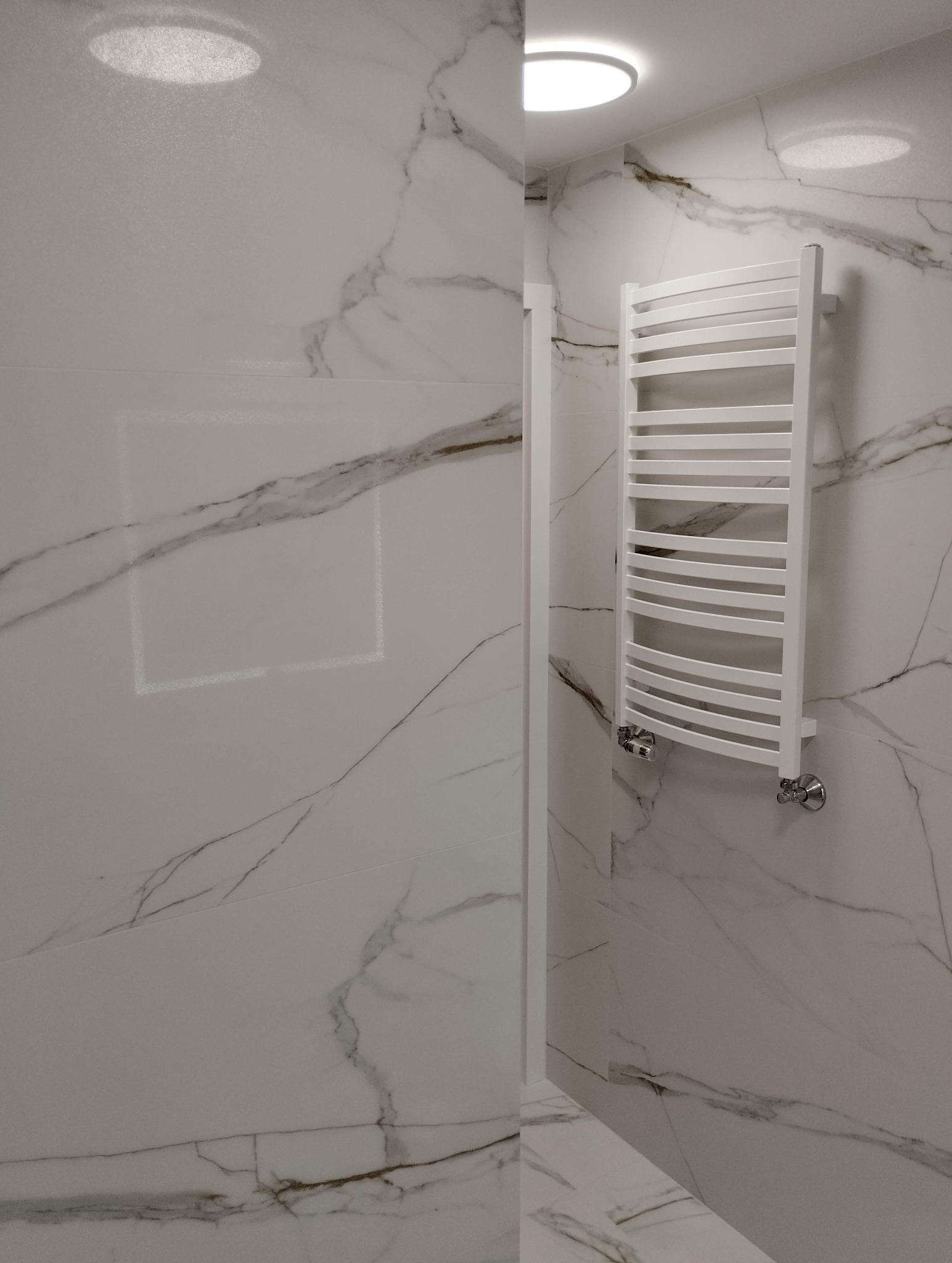 Biała łazienka z marmurowymi płytkami i grzejnikiem drabinkowym. Minimalistyczny design, oświetlenie sufitowe. Widok na kabinę prysznicową.