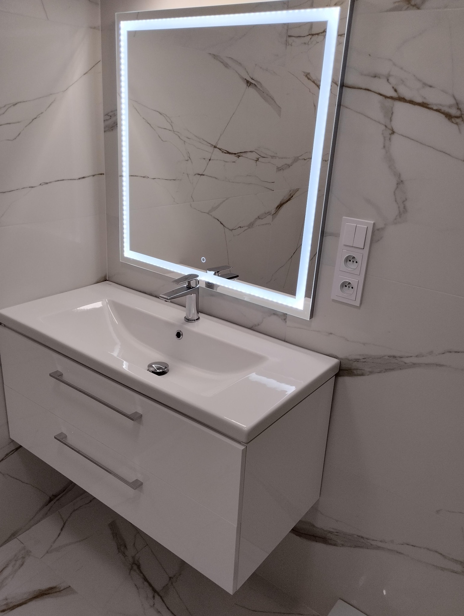 Biała umywalka z szafką i podświetlanym lustrem na tle marmurowych płytek w łazience. Nowoczesny design, chromowana bateria, gniazdka elektryczne.
