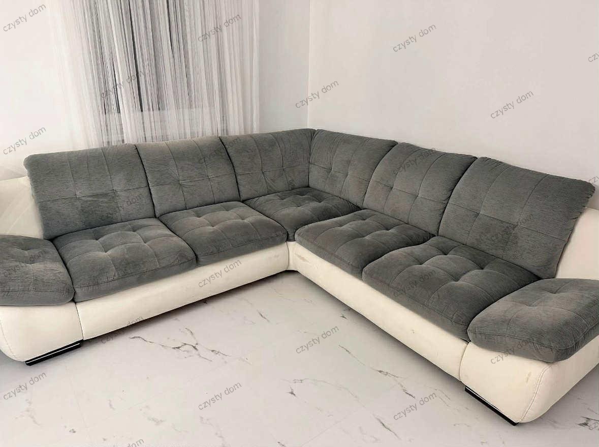 Szara, narożna kanapa z białymi bokami na marmurowej podłodze. Widoczne poduszki z kwadratowymi przeszyciami i zasłony w tle. Ujęcie z niskiego kąta.