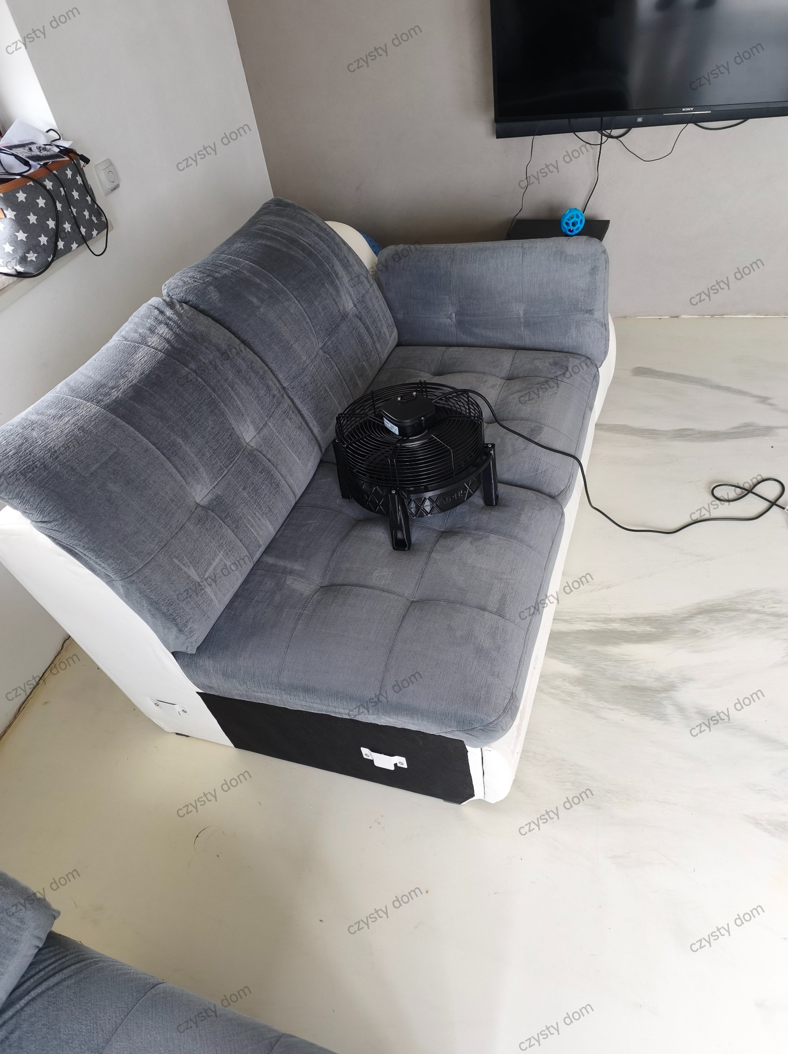 Szara, modułowa kanapa z wentylatorem na siedzisku, ustawiona w rogu pokoju z telewizorem. Widoczne ślady użytkowania na podłodze.