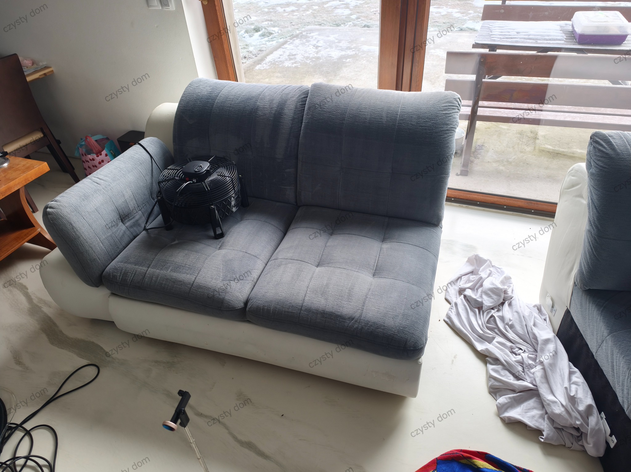 Szara kanapa z wentylatorem obok okna. Na podłodze widoczny sprzęt do czyszczenia i biała tkanina. Ujęcie z góry, wnętrze domu.