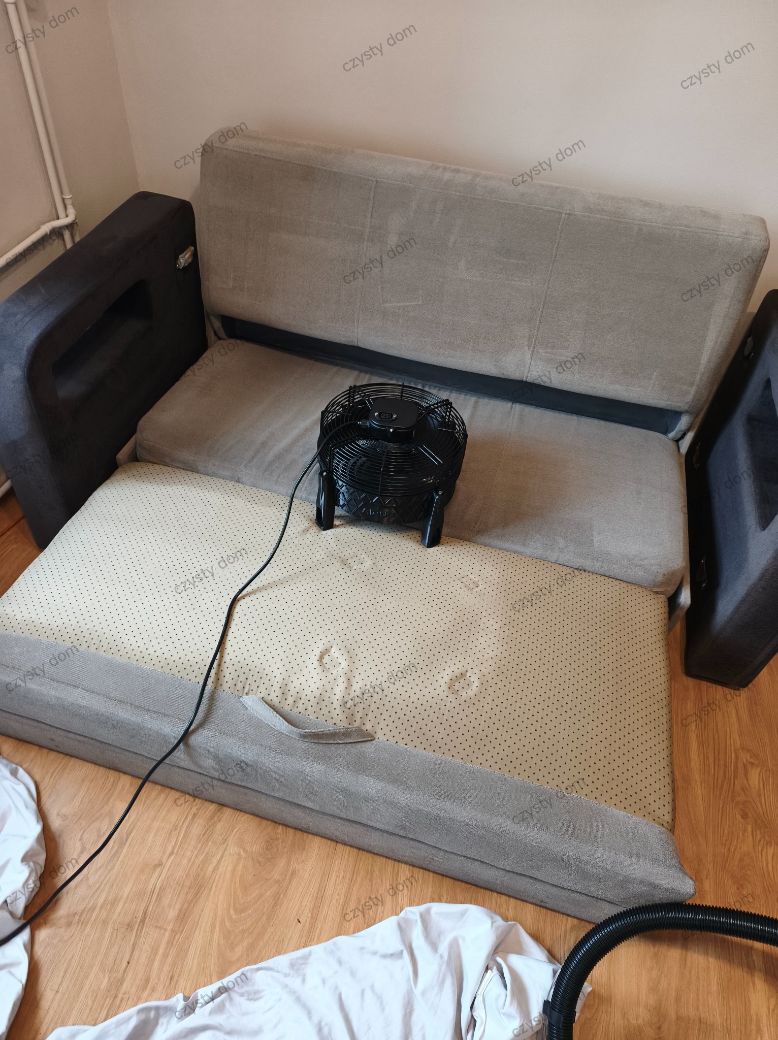 Rozłożona kanapa z wentylatorem i wężem odkurzacza. Widoczne ślady po czyszczeniu na tapicerce. Przygotowanie do suszenia po praniu.