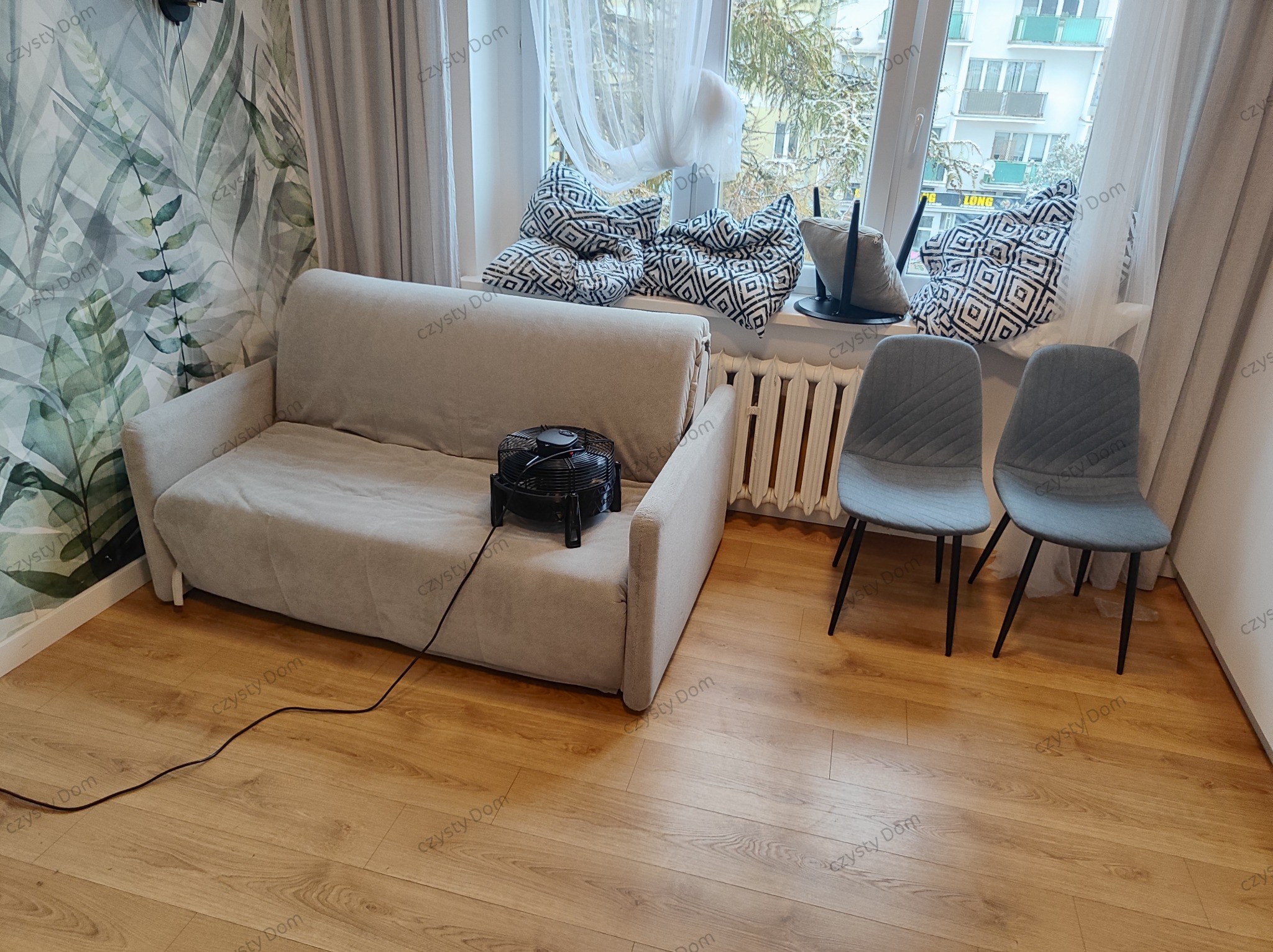 Jasna kanapa z wentylatorem na siedzisku, dwie szare krzesła przy oknie z poduszkami. Parkiet na podłodze i tapeta w liście na ścianie.