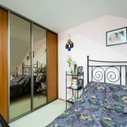 Szafa z przesuwnymi, lustrzanymi drzwiami w sypialni na poddaszu, odbijająca łóżko z metalową ramą i stolik z dekoracjami.