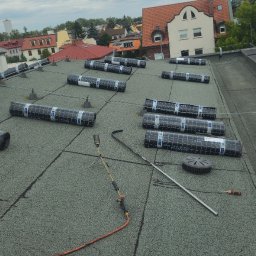 DACH SERWIS Łukasz Lewicki - Montaż papy termozgrzewalnej na płaskim dachu. Widoczne rolki papy Bikutop, palnik dekarski i narzędzia. W tle zabudowa miejska i kominy przemysłowe.