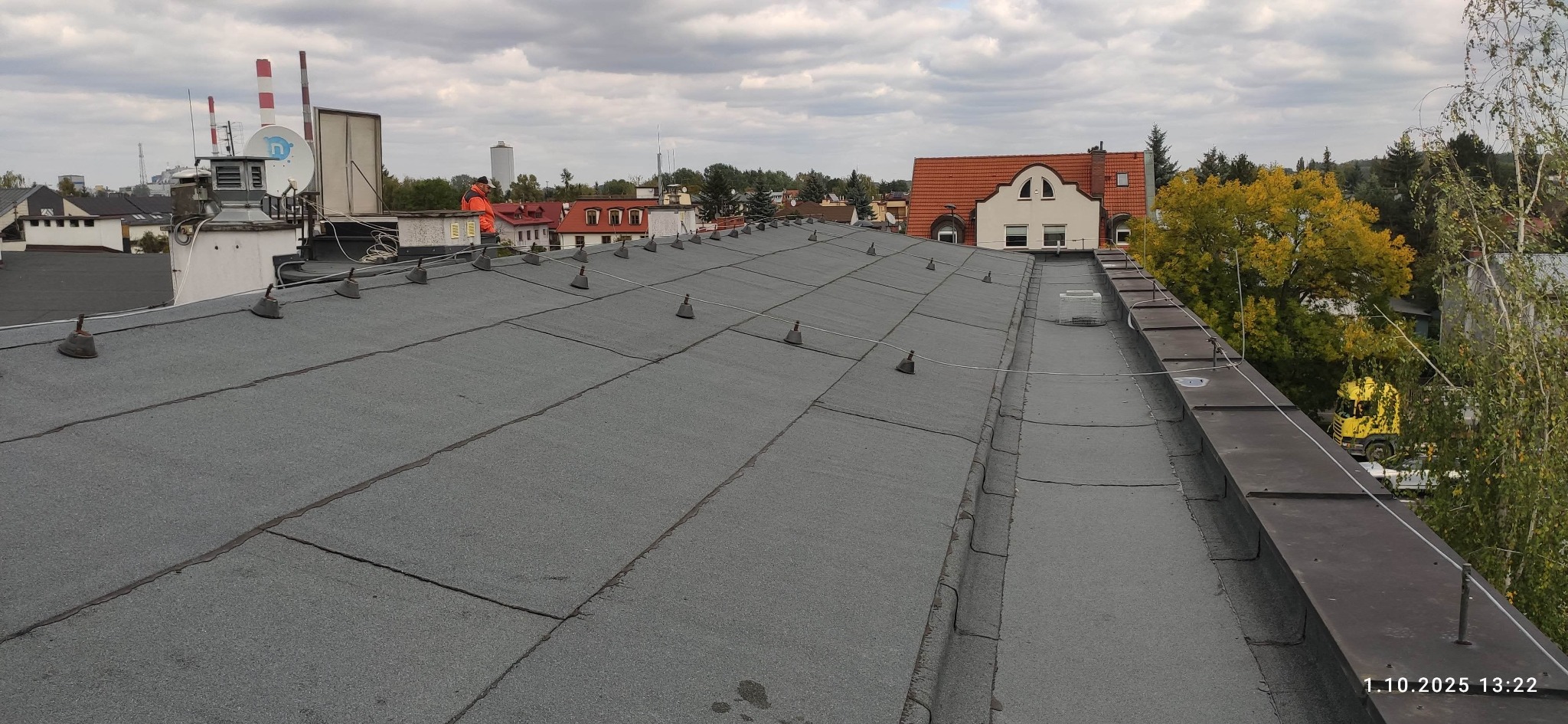 Szary dach pokryty papą z odgromnikami na tle zabudowań miejskich i pochmurnego nieba. Widoczny komin i drzewa. Ujęcie z góry.