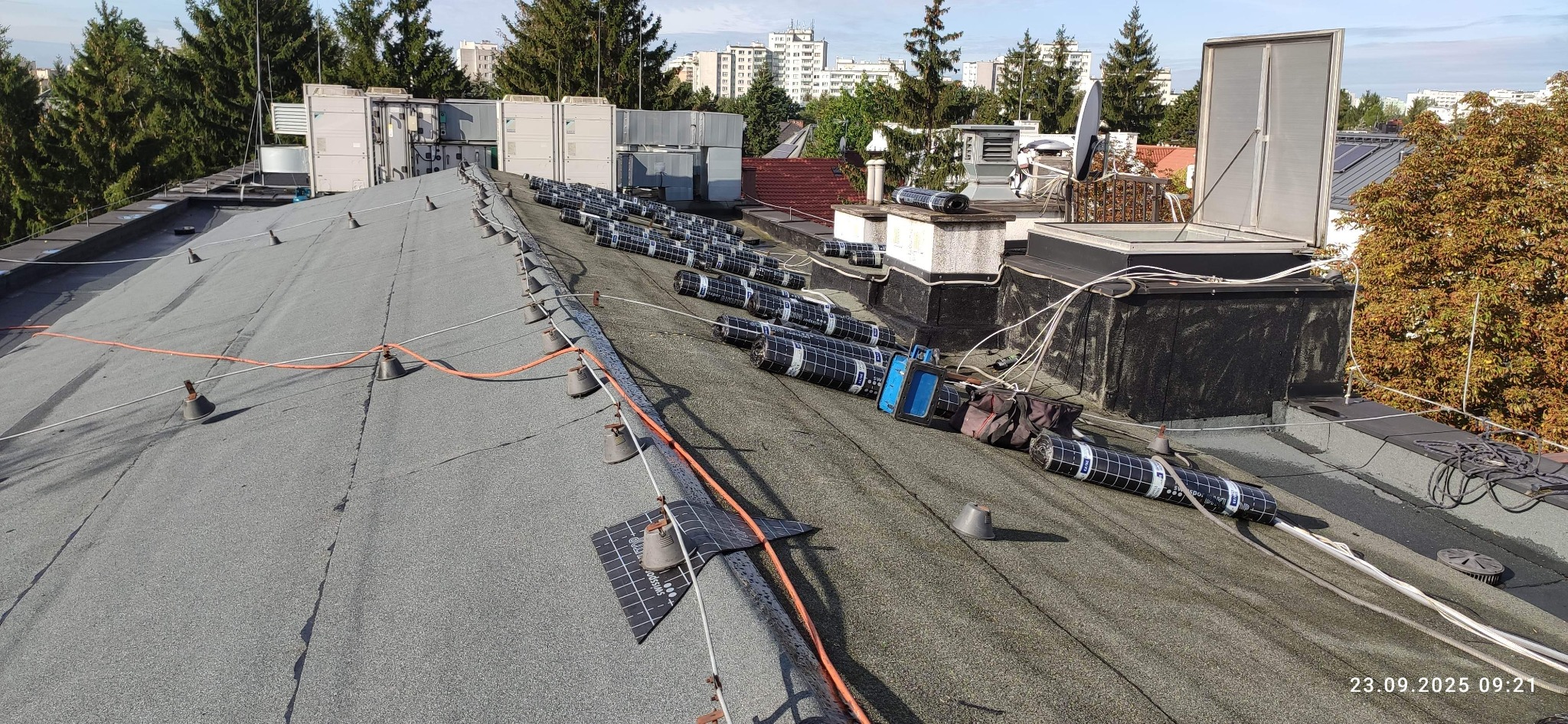 Dach pokryty papą z przygotowanymi rolkami izolacji termicznej, widoczne elementy wentylacji i instalacji odgromowej. Prace dekarskie w toku, widok z góry.