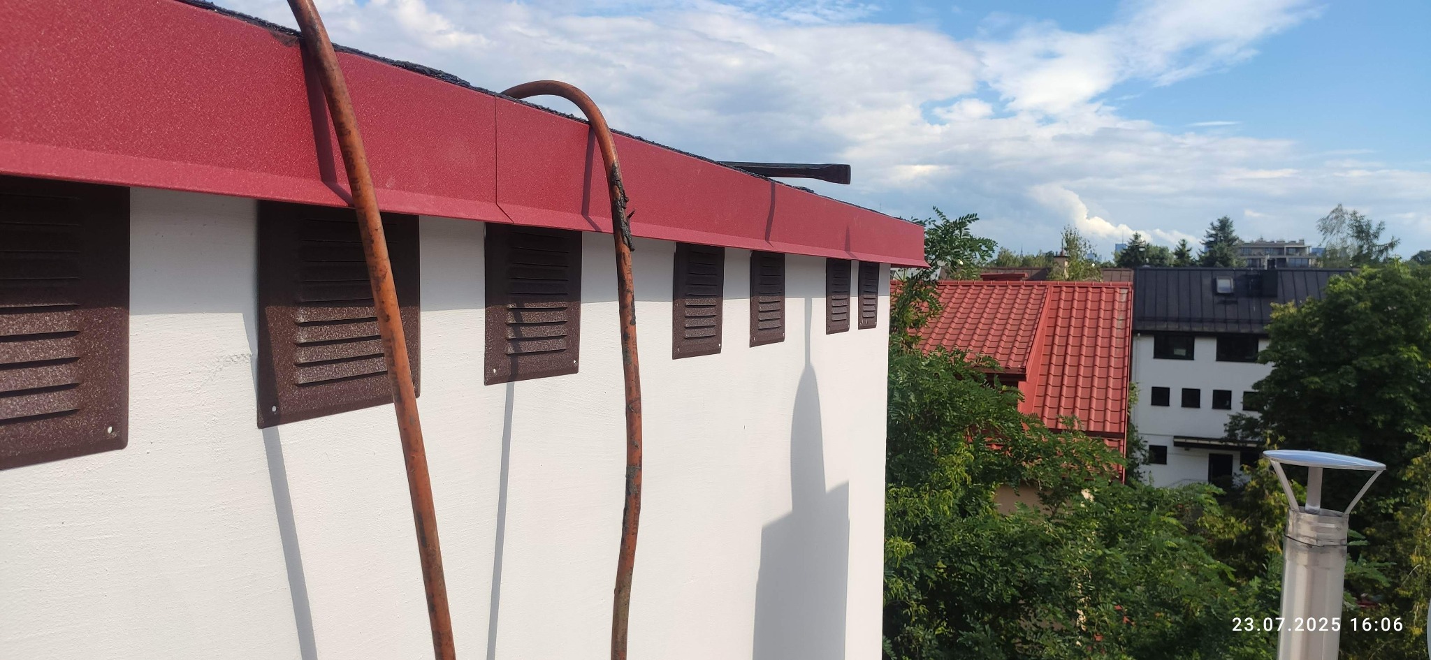 Fragment dachu z czerwonym pokryciem i białej ściany z brązowymi otworami wentylacyjnymi. W tle zieleń drzew i budynki. Widoczny przewód. Ujęcie z dołu.