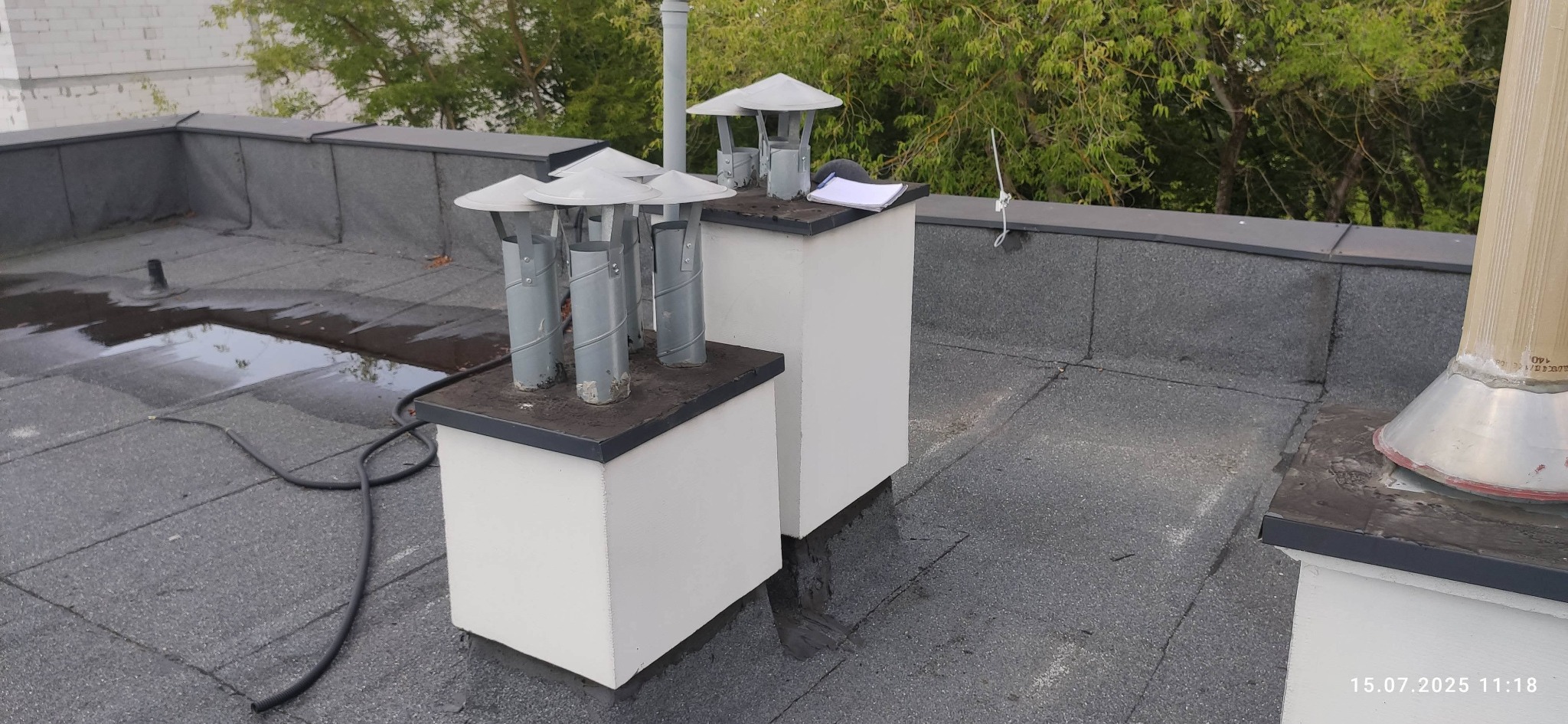 Dach pokryty papą z kominami wentylacyjnymi i fragmentem instalacji odgromowej. Na kominie leży notatnik. Widoczne ślady wilgoci na dachu.