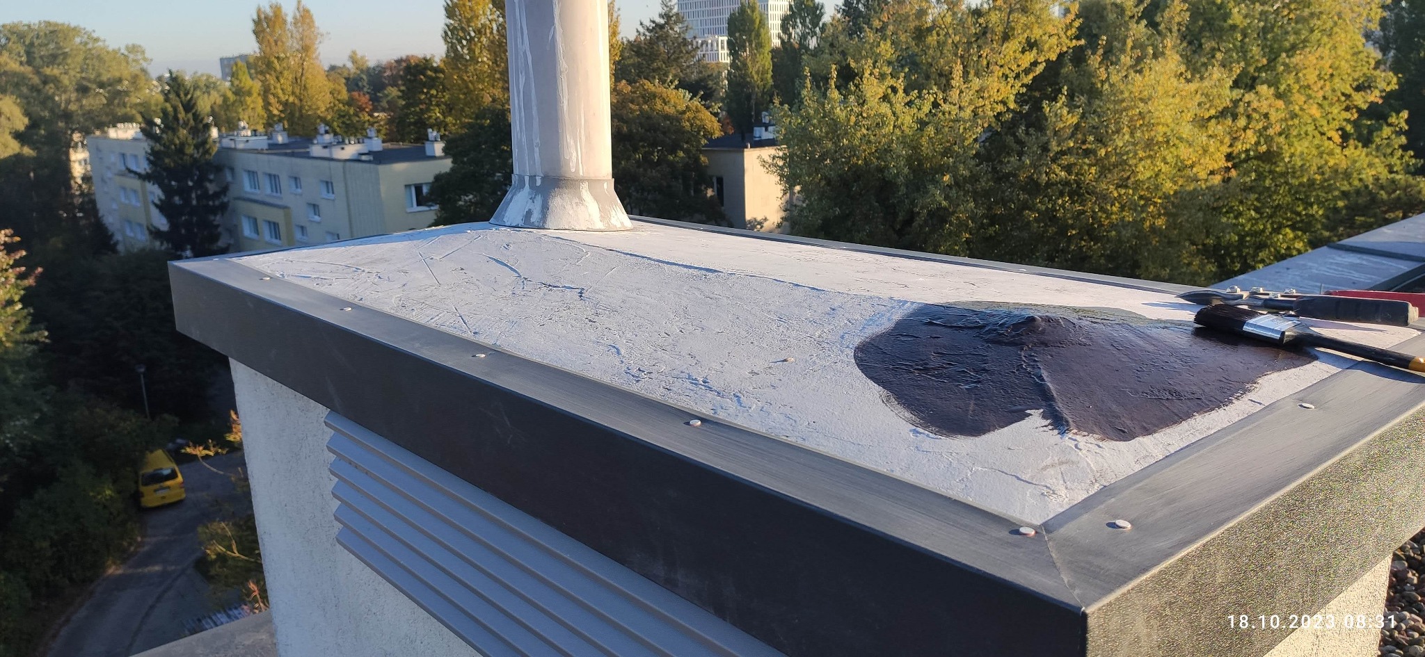 Biały dach pokryty częściowo ciemną farbą, obok pędzle i narzędzia. Widok z góry na tle zieleni i budynków. Etap prac malarskich na dachu płaskim.