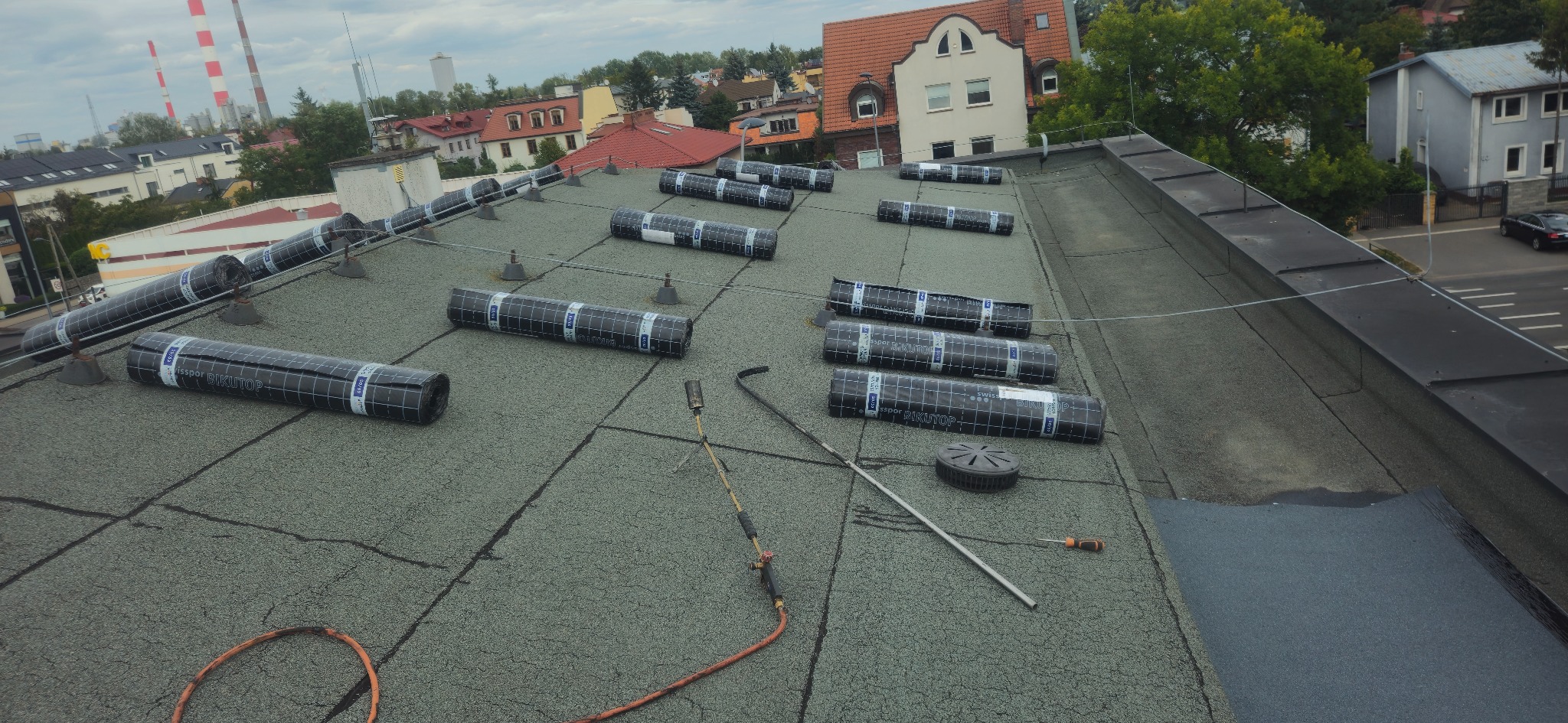 Montaż papy termozgrzewalnej na płaskim dachu. Widoczne rolki papy Bikutop, palnik dekarski i narzędzia. W tle zabudowa miejska i kominy przemysłowe.