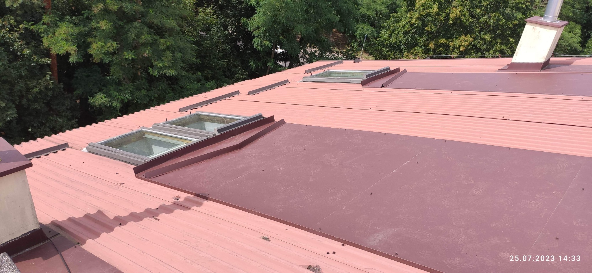 Czerwony dach z blachy trapezowej z oknami dachowymi i obróbkami blacharskimi, widok z góry w słoneczny dzień. Widoczne ślady stóp na panelu.