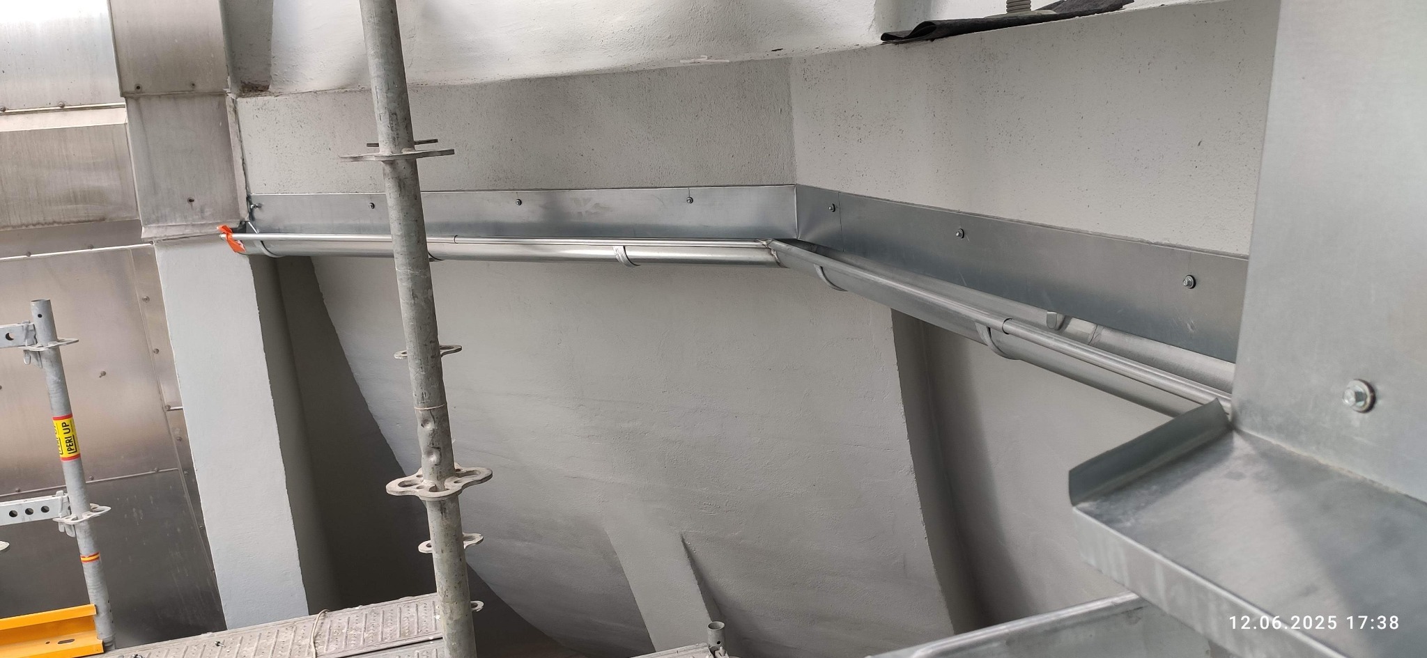 Zamontowane, metalowe rynny w narożniku budynku, widoczne z rusztowania. Rynny przymocowane do ściany za pomocą śrub. Jasne, neutralne tło.