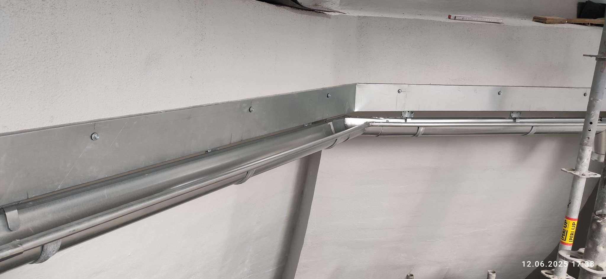 Zamontowana, metalowa rynna narożna wewnątrz budynku, przymocowana do ściany za pomocą śrub. Widoczne elementy rusztowania.