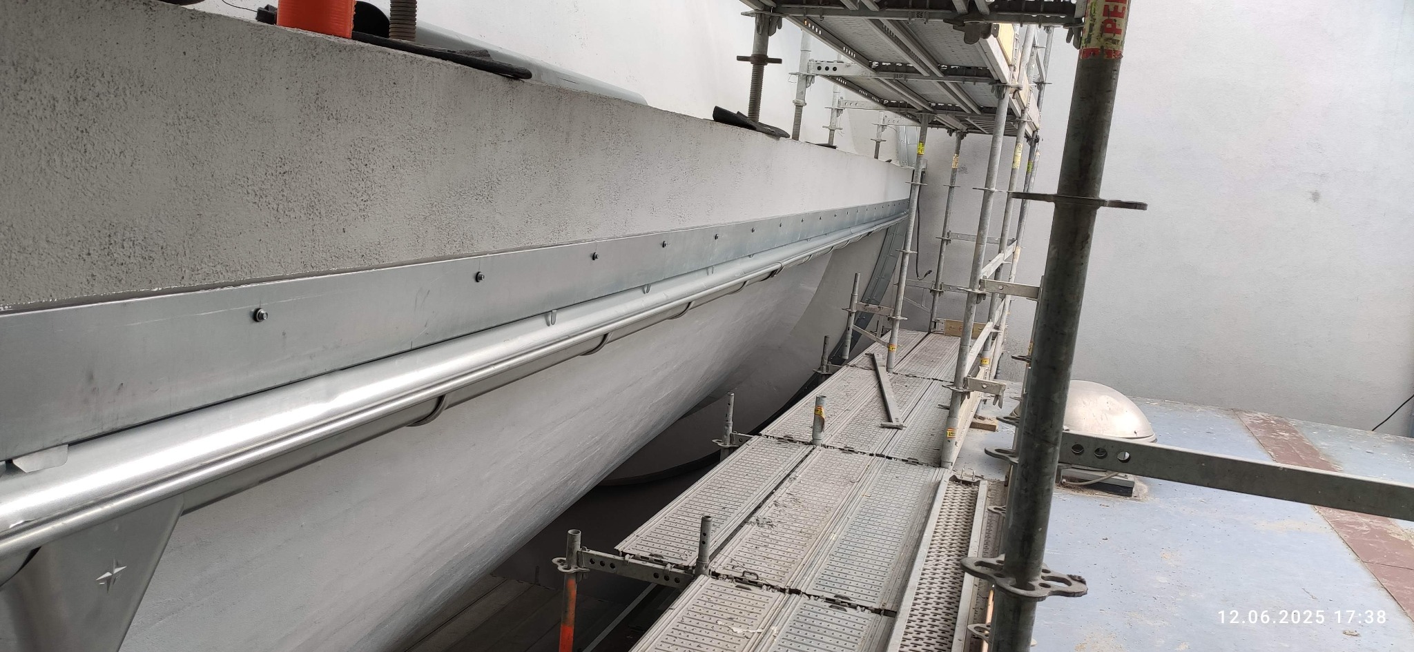 Szare rusztowanie przy ścianie z zamontowaną metalową rynną. Widoczne śruby mocujące. Ujęcie z dołu, podkreślające detale instalacji.