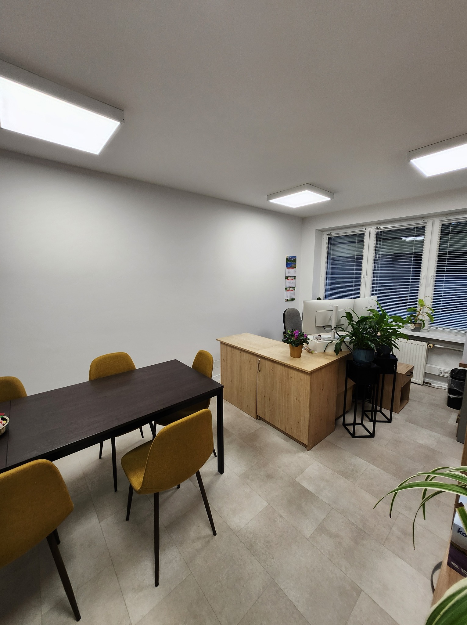 Jasne biuro z drewnianym biurkiem, dwoma monitorami, roślinami doniczkowymi i stołem konferencyjnym z żółtymi krzesłami. Minimalistyczny wystrój, neutralna kolorystyka.