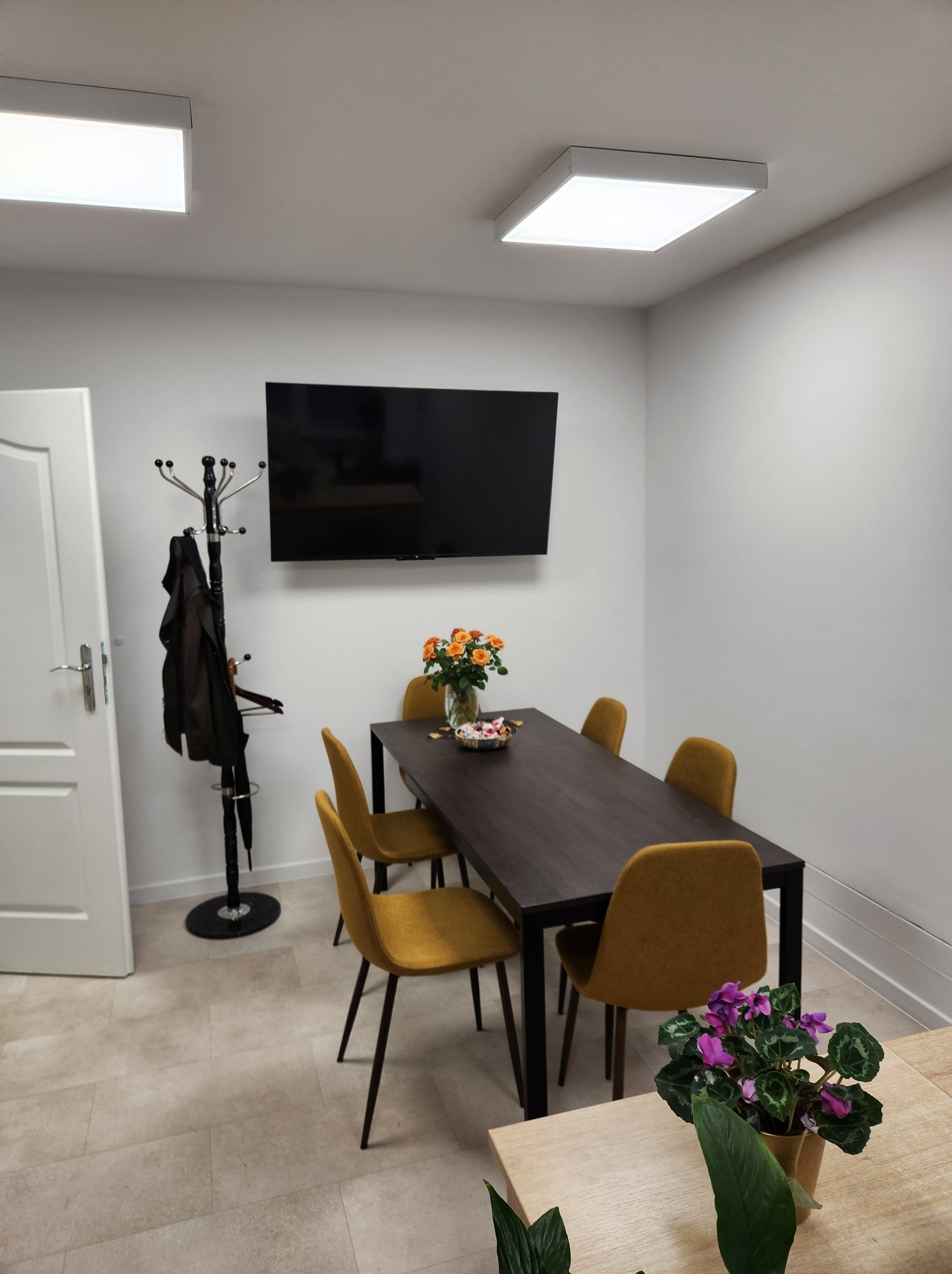 Jasne biuro z ciemnym stołem, żółtymi krzesłami i telewizorem na ścianie. Na stole wazon z kwiatami i miseczka. W rogu wieszak na ubrania i drzwi.