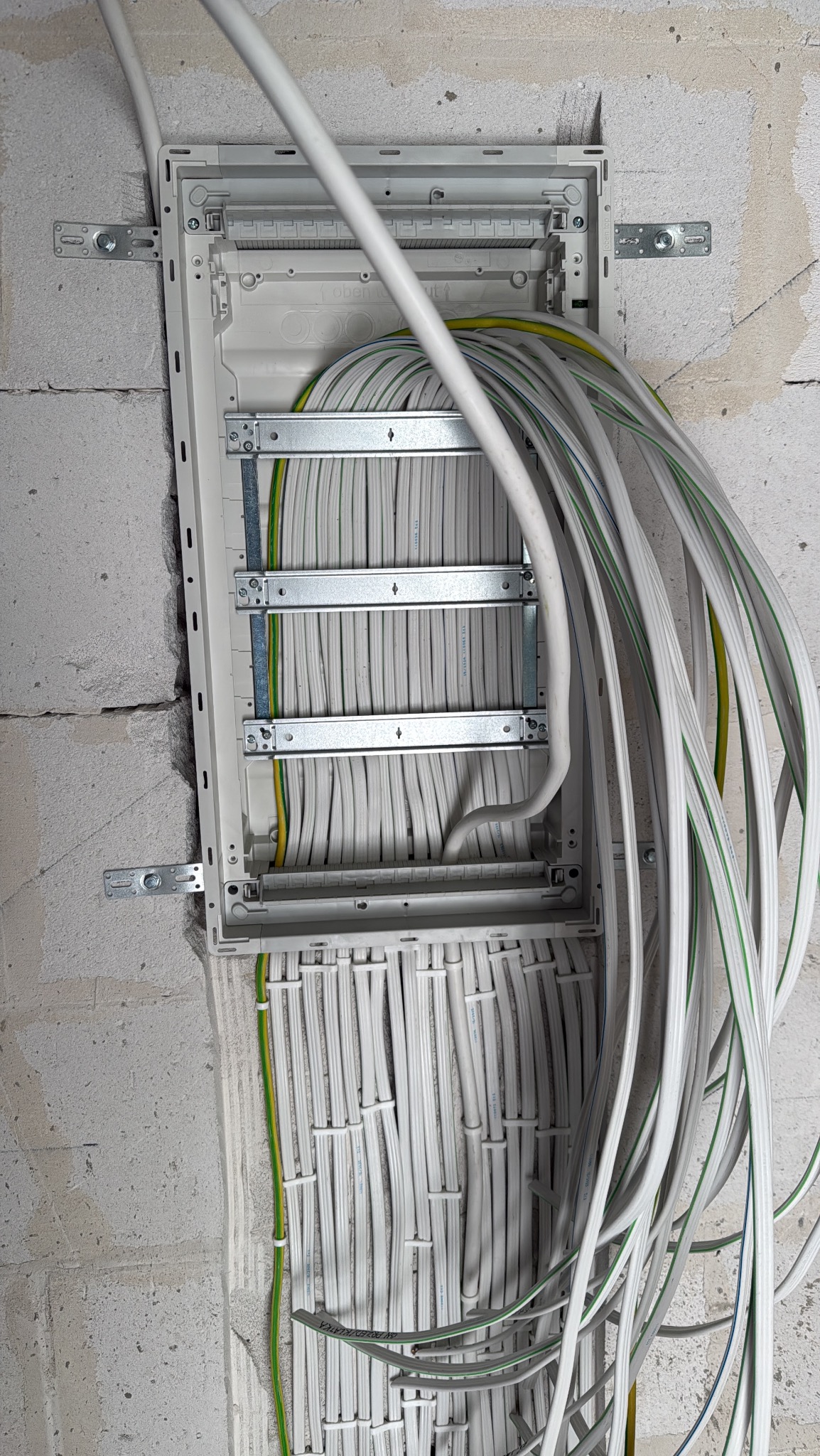 Rozdzielnica elektryczna z licznymi przewodami wewnątrz, zamontowana na surowej ścianie z betonu komórkowego. Widoczne metalowe uchwyty i uporządkowane kable.