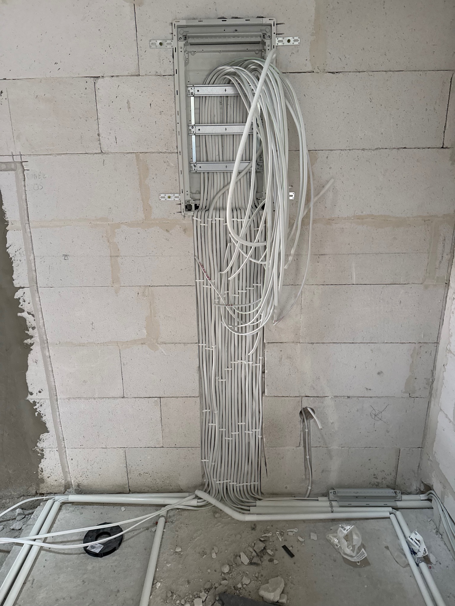 Rozdzielnia elektryczna z licznymi przewodami w białej osłonie, zamontowana na surowej ścianie z betonu komórkowego. Widoczne rury instalacyjne przy podłodze.