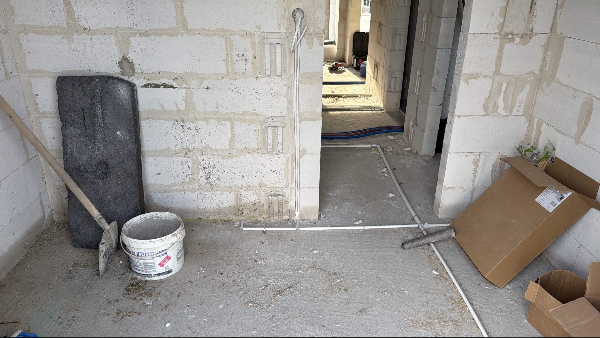 Surowy stan: Instalacja elektryczna w bruzdach ściany z betonu komórkowego, rury w podłodze, narzędzia i kartony. Widok na kolejne pomieszczenie z instalacjami.