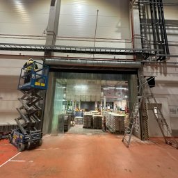 MiM Krystian Rumiński - Montaż bramy przemysłowej szybkobieżnej w hali magazynowej. Pracownik na podnośniku nożycowym, widoczna drabina i palety z towarem w tle.