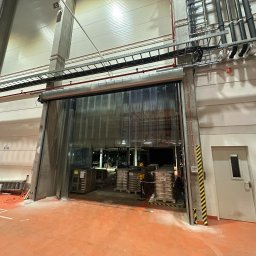 MiM Krystian Rumiński - Otwarta brama przemysłowa z przezroczystych pasów PCV w hali magazynowej. Widoczne instalacje pod sufitem i wyposażenie wnętrza hali.