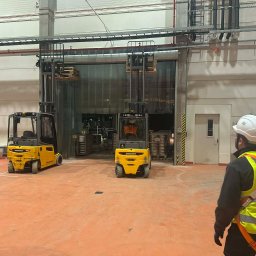 MiM Krystian Rumiński - Montaż bramy przemysłowej szybkobieżnej: widok na wózki widłowe i pracownika w kasku na tle otwartej bramy kurtynowej w hali magazynowej.